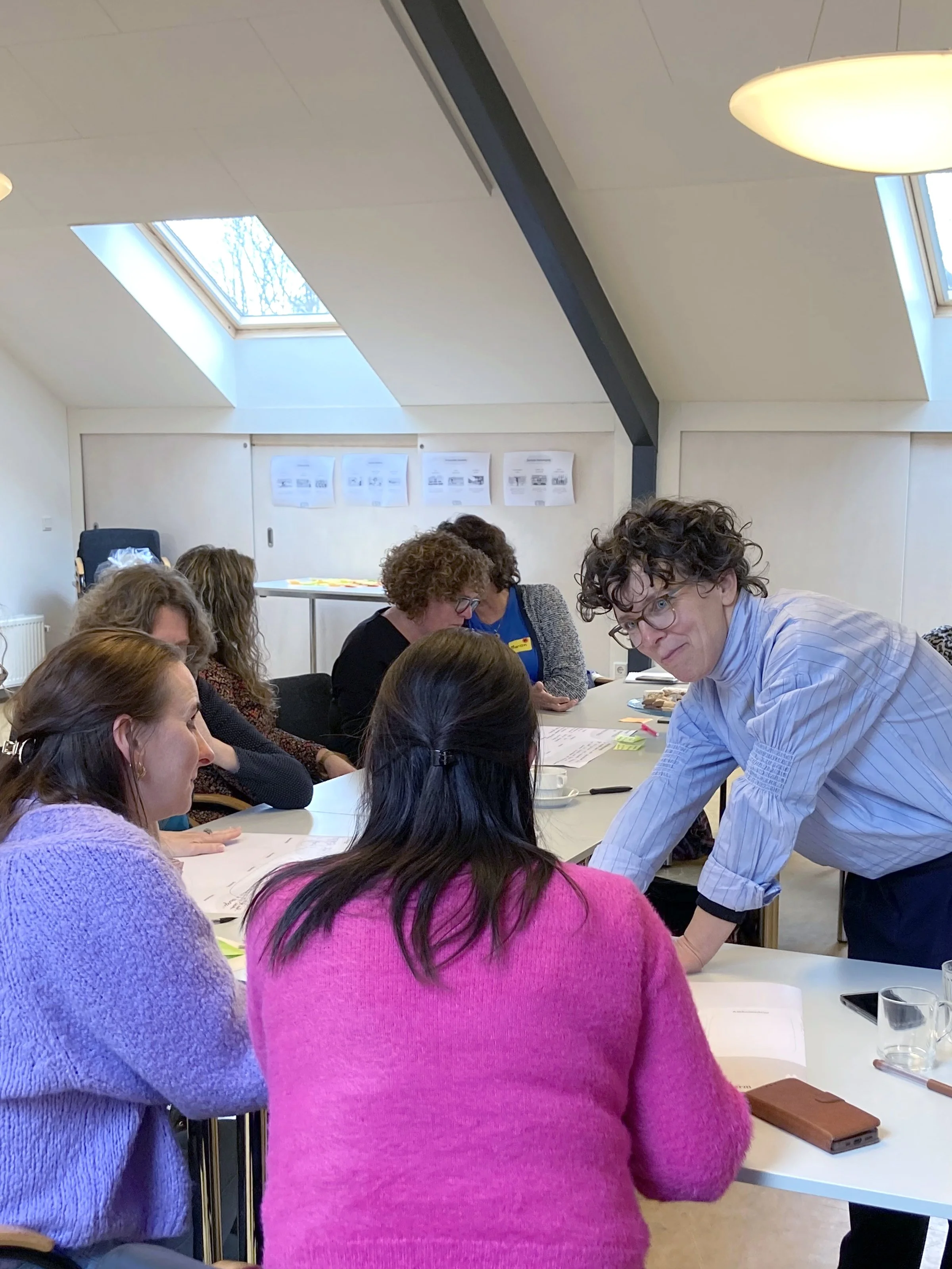 Groep mensen die in een kantoorruimte of vergaderruimte een bijeenkomst of training volgen, met een man die iets uitlegt aan een vrouw, terwijl andere mensen opletten, tijdens een workshop of vergadering in een kantoor met schuine plafonds en daglicht uit dakramen.