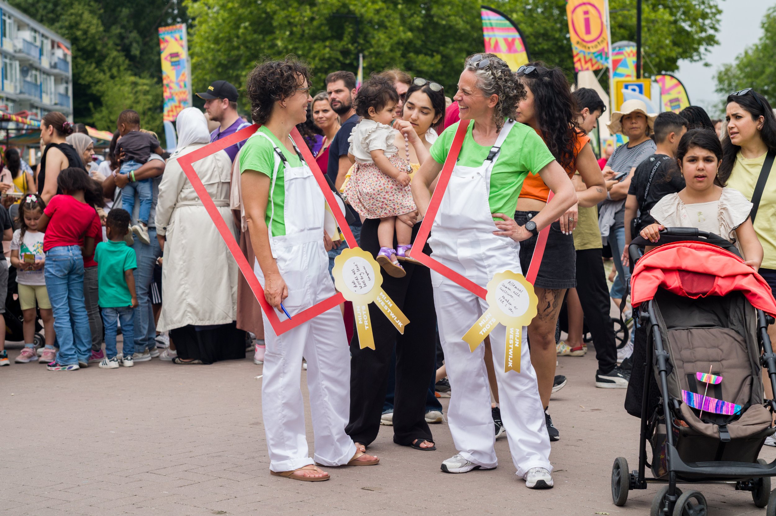 Vrouwencollege op een druk festival, twee vrouwen in witte kleren en groene shirts lachen en praten, terwijl ze grote rode fotolijsten vasthouden, tussen veel bezoekers, kinderen en een kinderwagen.