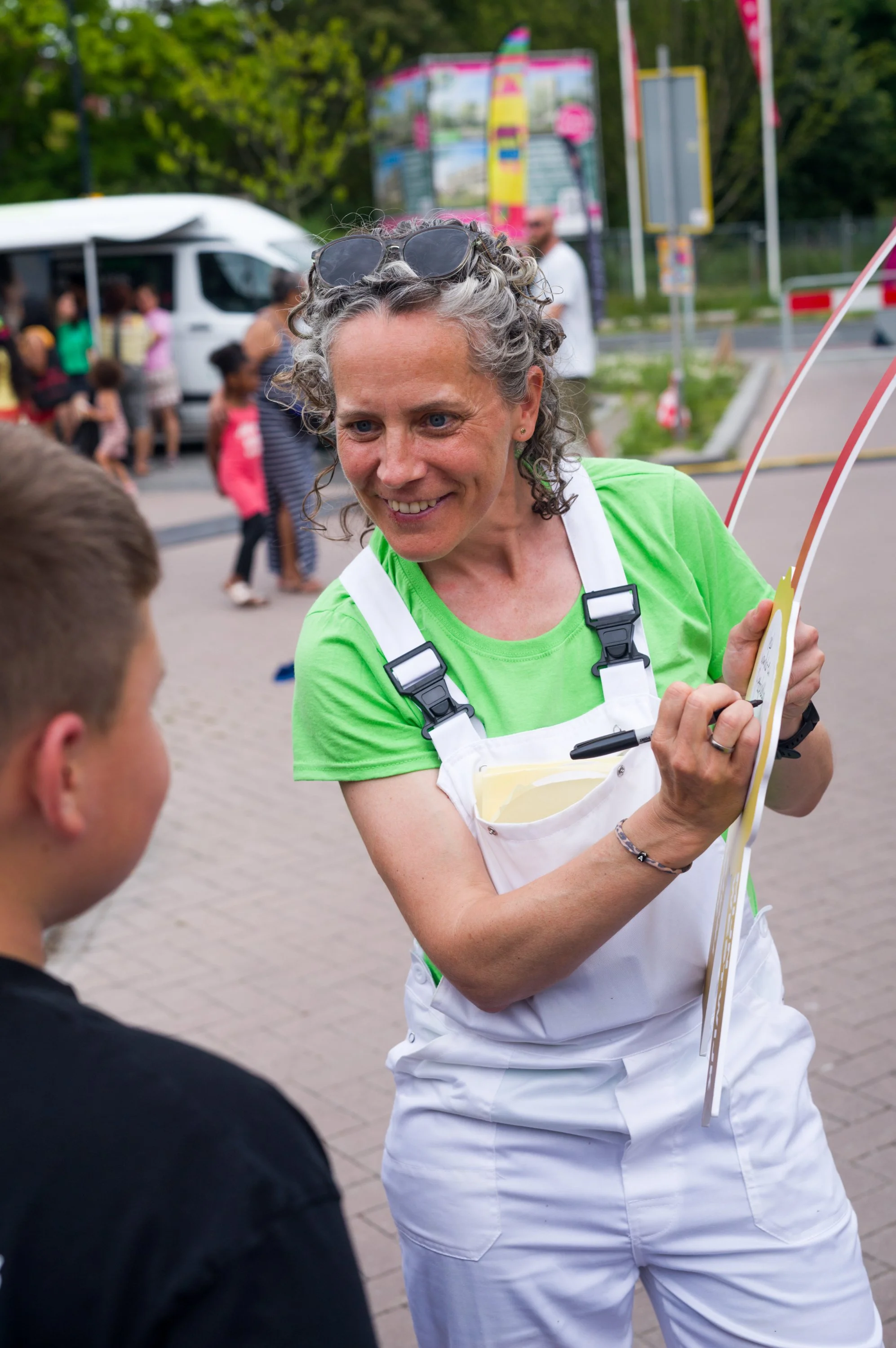 Vrouw met grijze, krullende haar en een groene T-shirt, glimlachend en met verstelbare brillen op haar hoofd, die een flipchart of bord vasthoudt en met een jonge jongen spreekt, op een drukke locatie met mensen op de achtergrond.