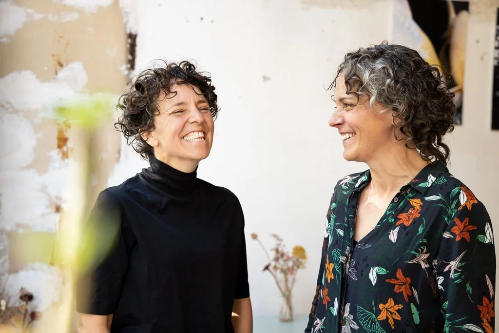 Twee vrouwen lachen naar elkaar in een kamer met een witte muur en een vaas met gedroogde bloemen op de achtergrond.