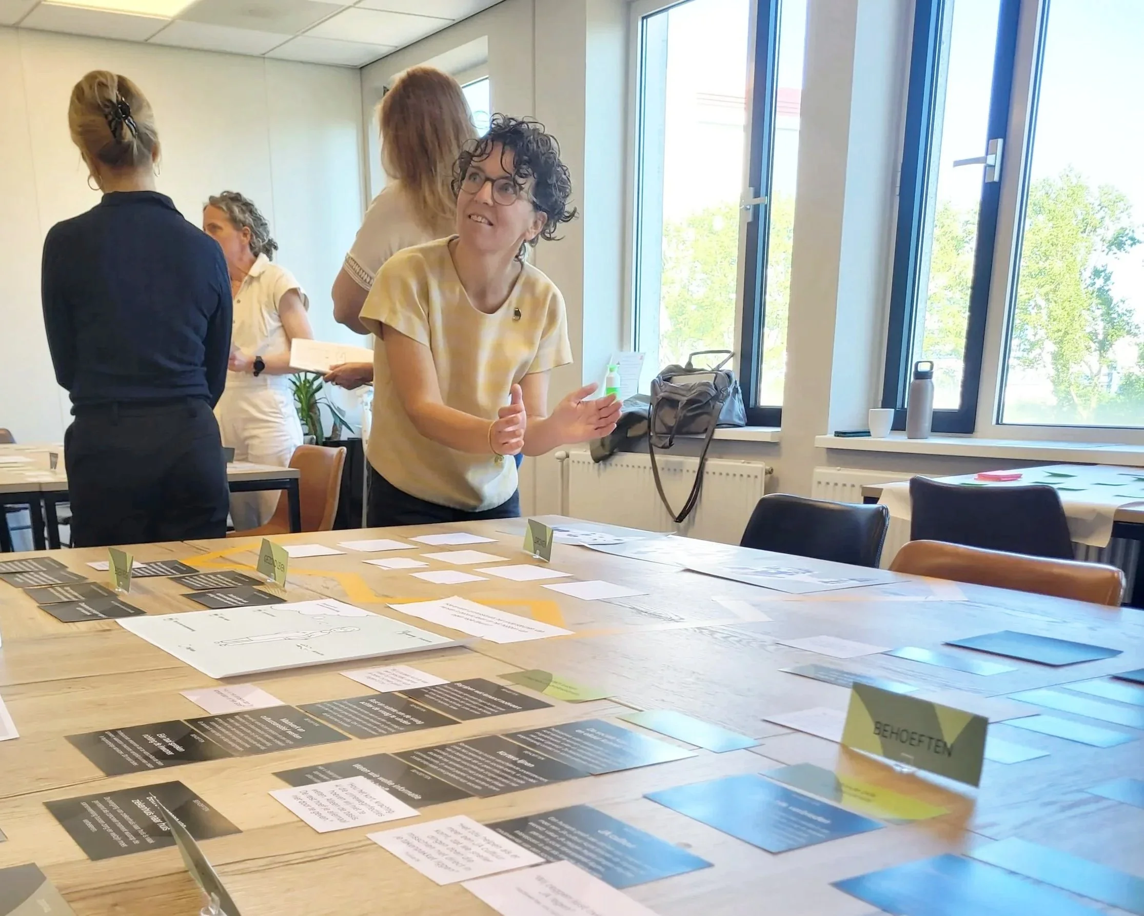 Vijftigers die in een vergaderkamer discussiëren, met een tafel vol informatiekaarten en documenten , grote ramen met uitzicht op bomen.