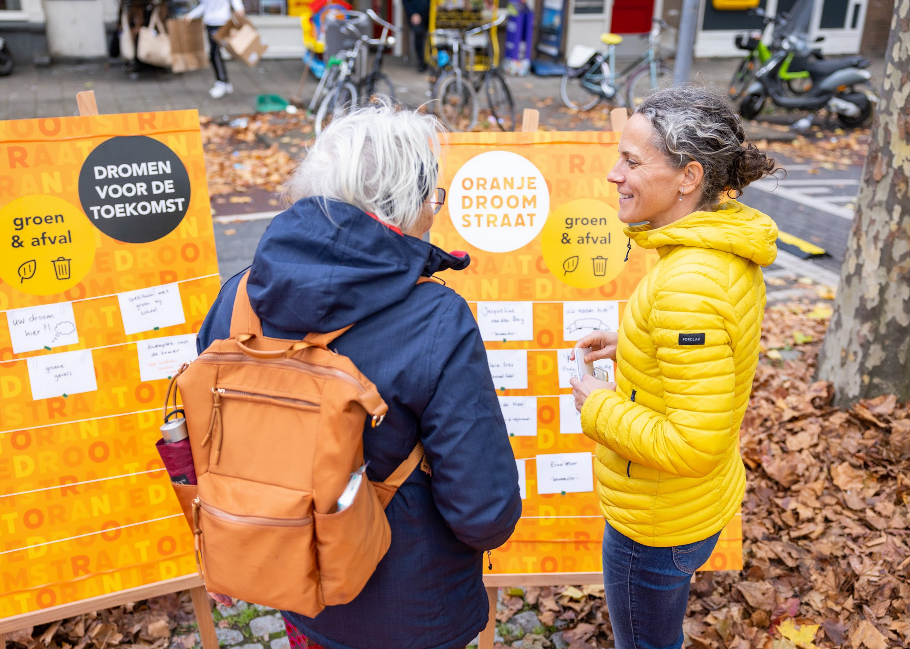 Twee vrouwen staan buiten op een straat met bomen en herfstbladeren, praten voor een oranje en witte prullenbak die is ingericht voor een milieuproject. Ze dragen jas en rugzak, en er staan fietsen en scooters op de achtergrond.