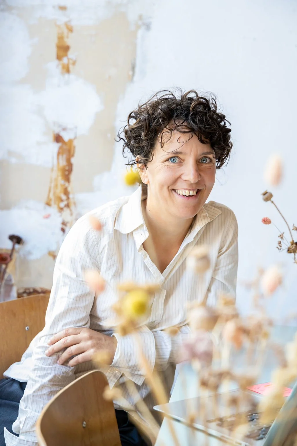 Vrouw met kort krullend haar en blauwe ogen die glimlacht, zittend aan een tafel met droogbloemen, tegen een muur met afwas en verfsporen.