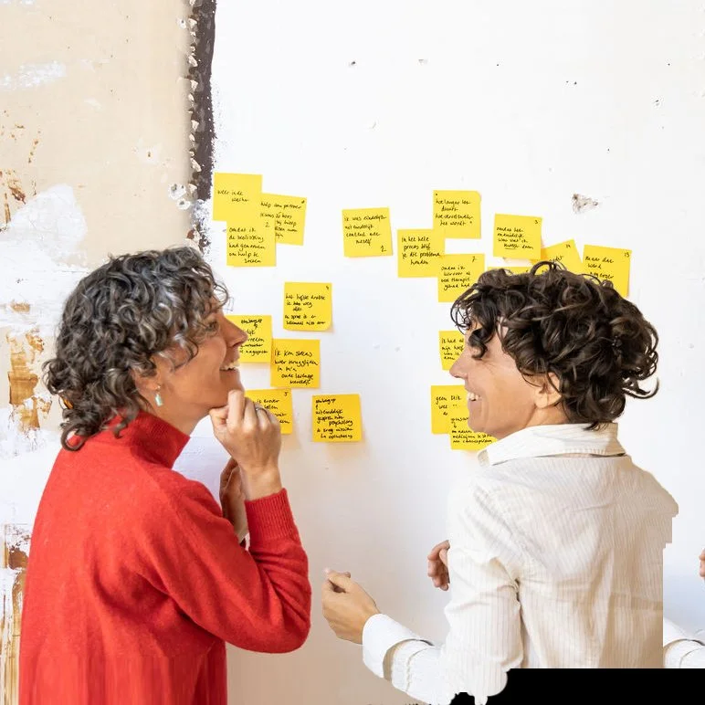 Twee vrouwen die lachen en praten bij een muur met gele notitiekaarten.