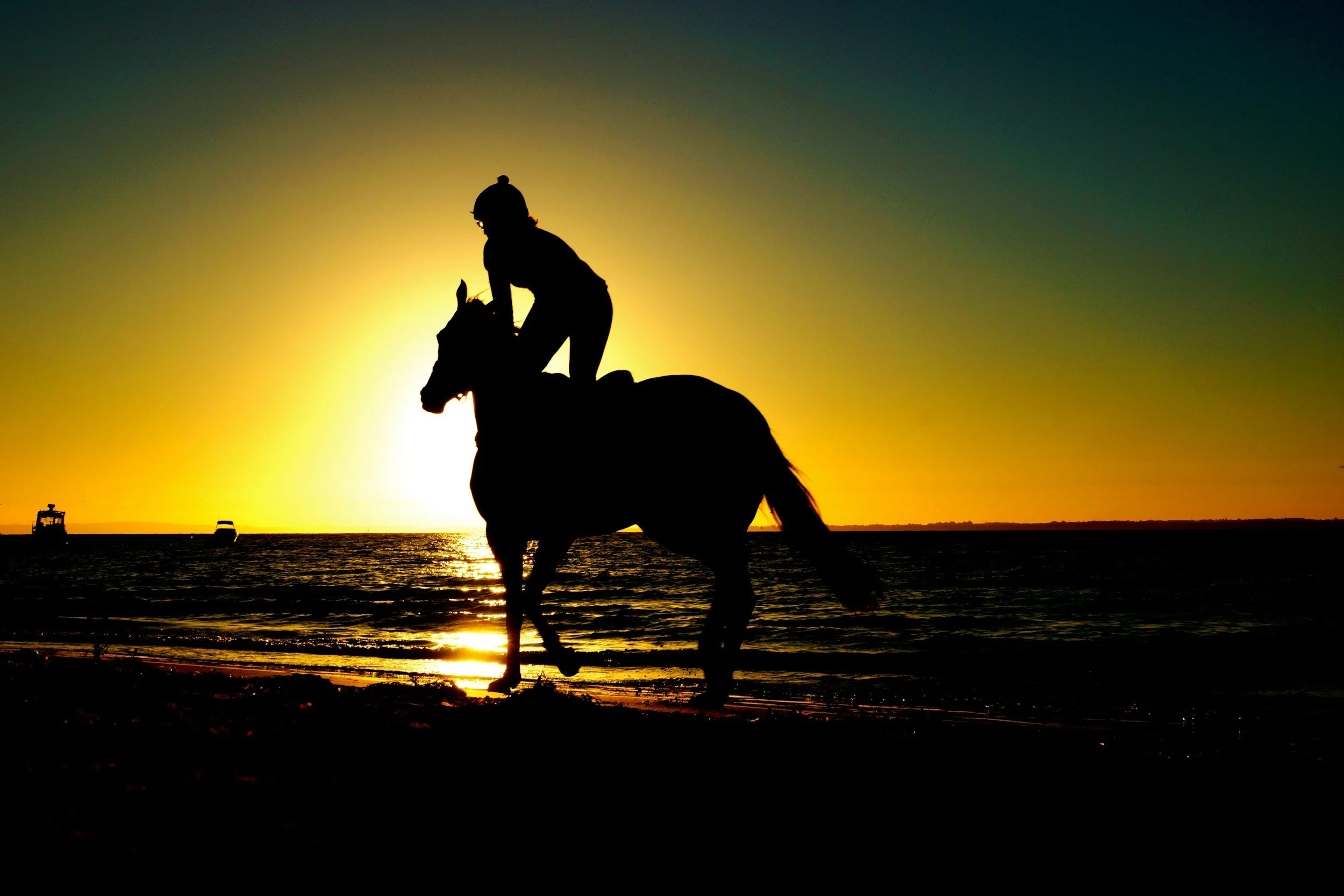 Silhouette of a person riding a horse on the beach during sunset, with boats in the water and a colorful sky.