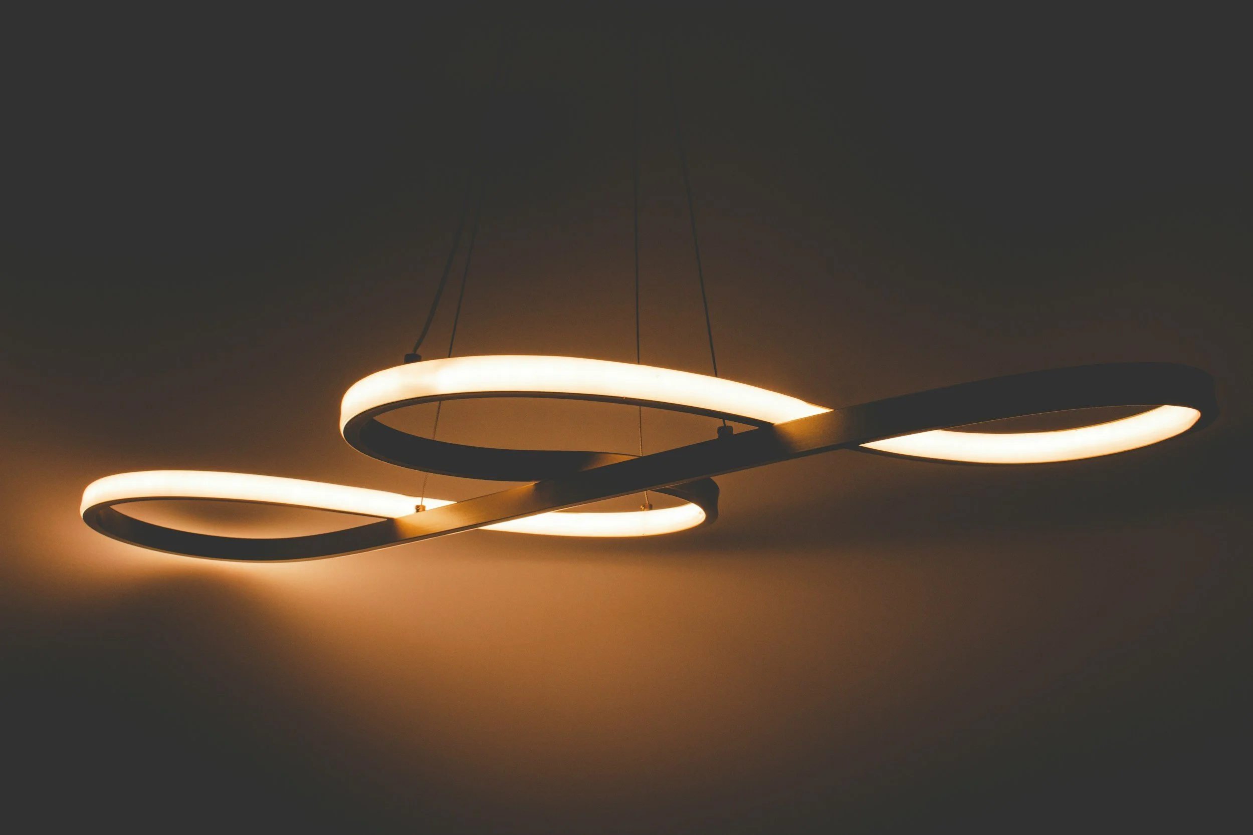 Modern circular pendant light fixture with a warm glow, suspended from the ceiling by thin wires, against a dark background.