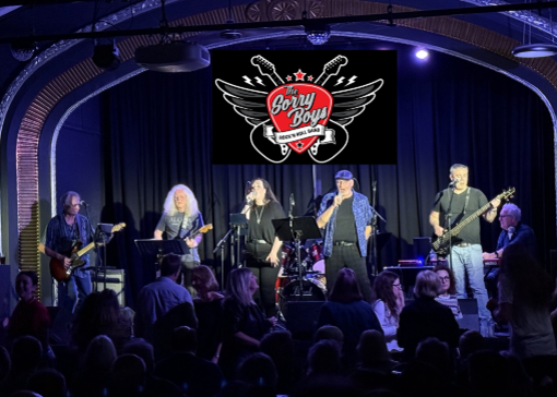A band performing on stage at a concert with a logo that says 'Gerry Boy's' with guitars and a singer in the center, audience watching in a dark indoor venue.