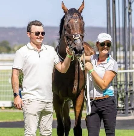 Cody Reardon standing outdoors with a dark bay thoroughbred, holding the horse on a lead during a professional racetrack or training‑day setting.