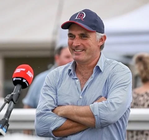 Tom Dabernig standing outdoors in a blue striped shirt and navy cap, speaking into a microphone during a professional racing interview.