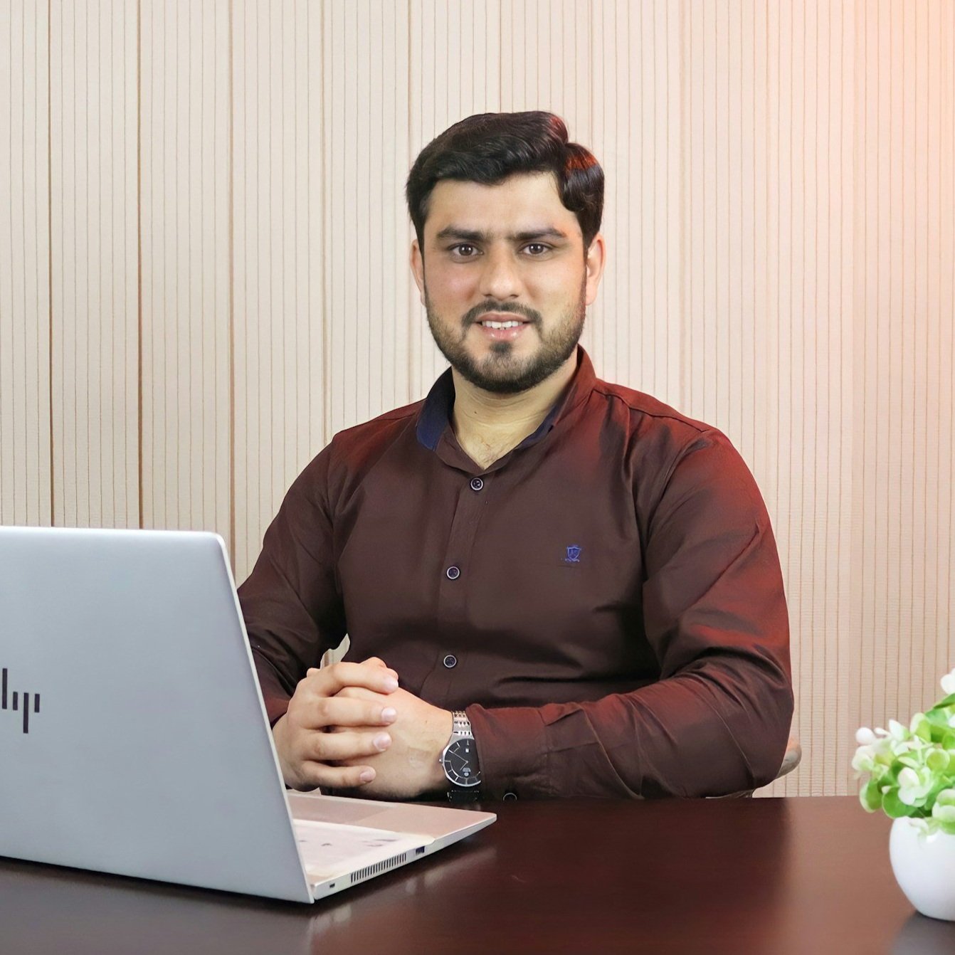 a man with dark hair and a beard sits at a desk with a laptop on it