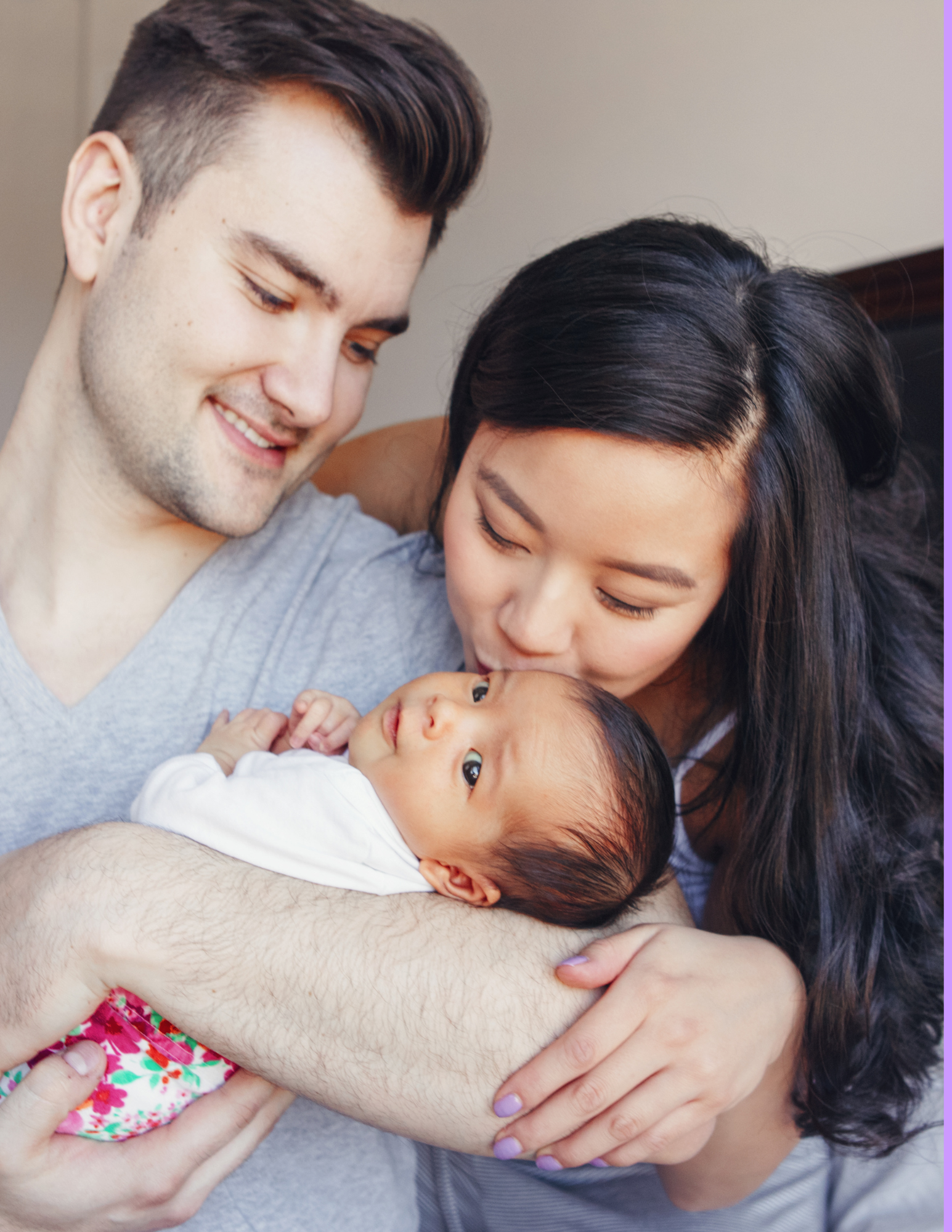A happy couple with a newborn baby, the woman and man smiling and leaning in close to the baby, who is lying on the man's arm, looking up