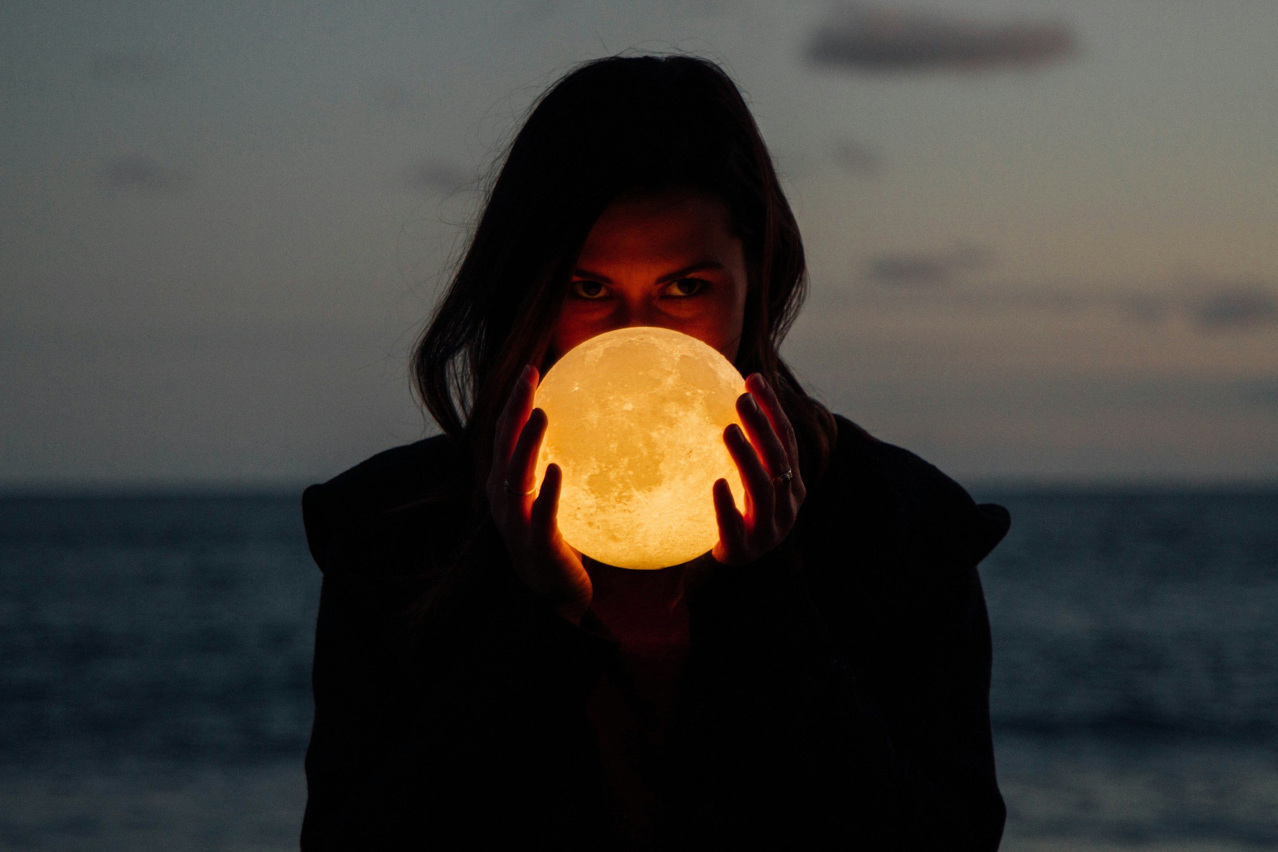 A person holding a glowing moon lamp at night outdoors with a cloudy sky and a body of water in the background.