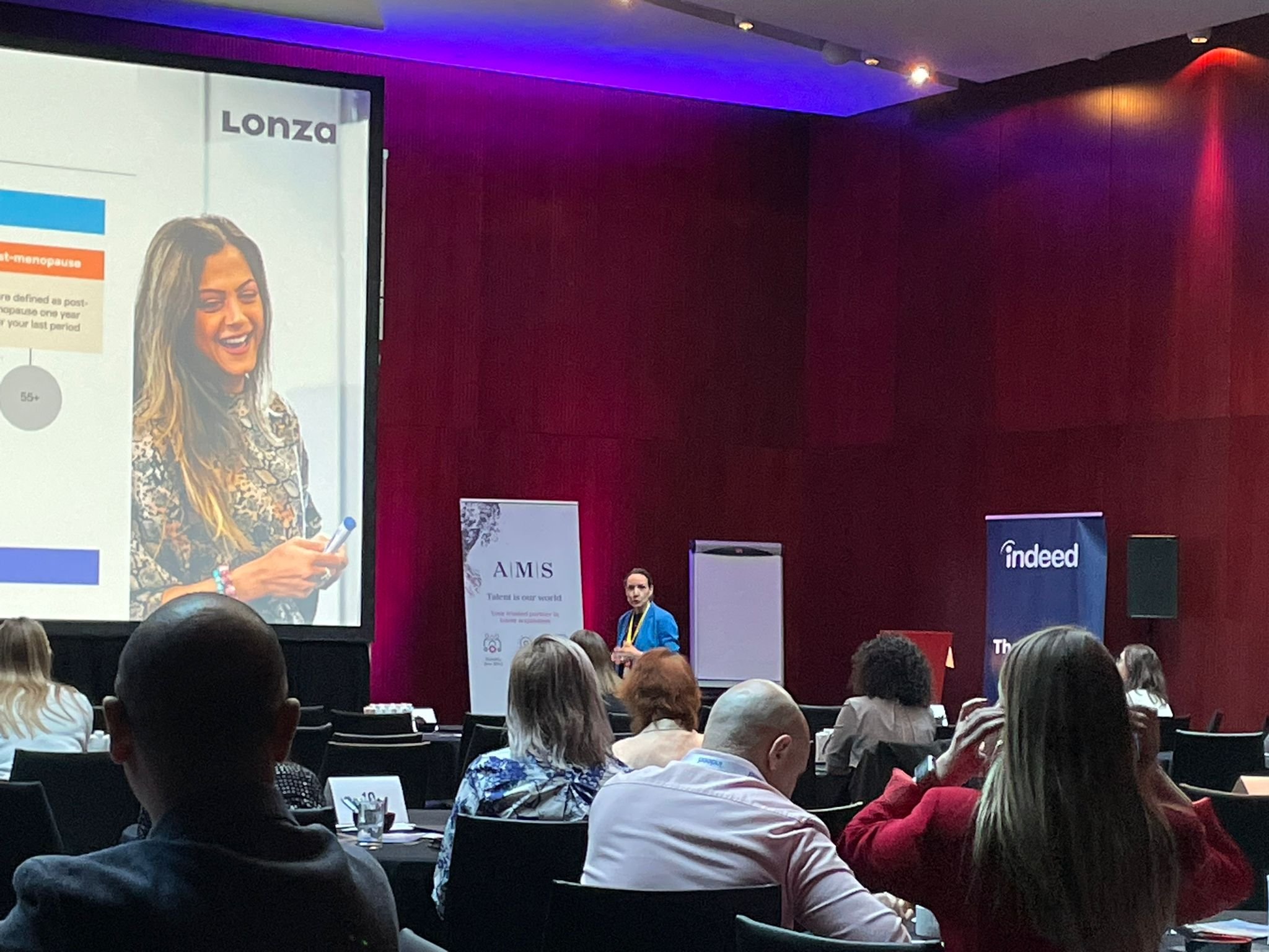 A conference room filled with attendees sitting at round tables, facing a presenter on stage. The presenter is a woman in a blue blazer standing near a whiteboard, with a large screen displaying her image and presentation on the left side. The background of the room has dark red walls and is illuminated with purple and red lighting. There are two banners, one for 'A M S' and another for 'Indeed,' near the stage.