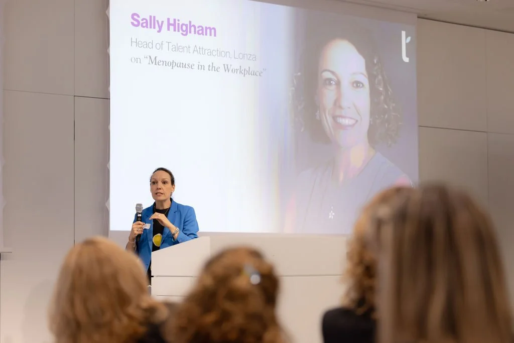 A woman in a blue blazer giving a presentation at a conference, with a large screen behind her displaying her photo and her name, Sally Higham, along with her title. Several audience members are listening.