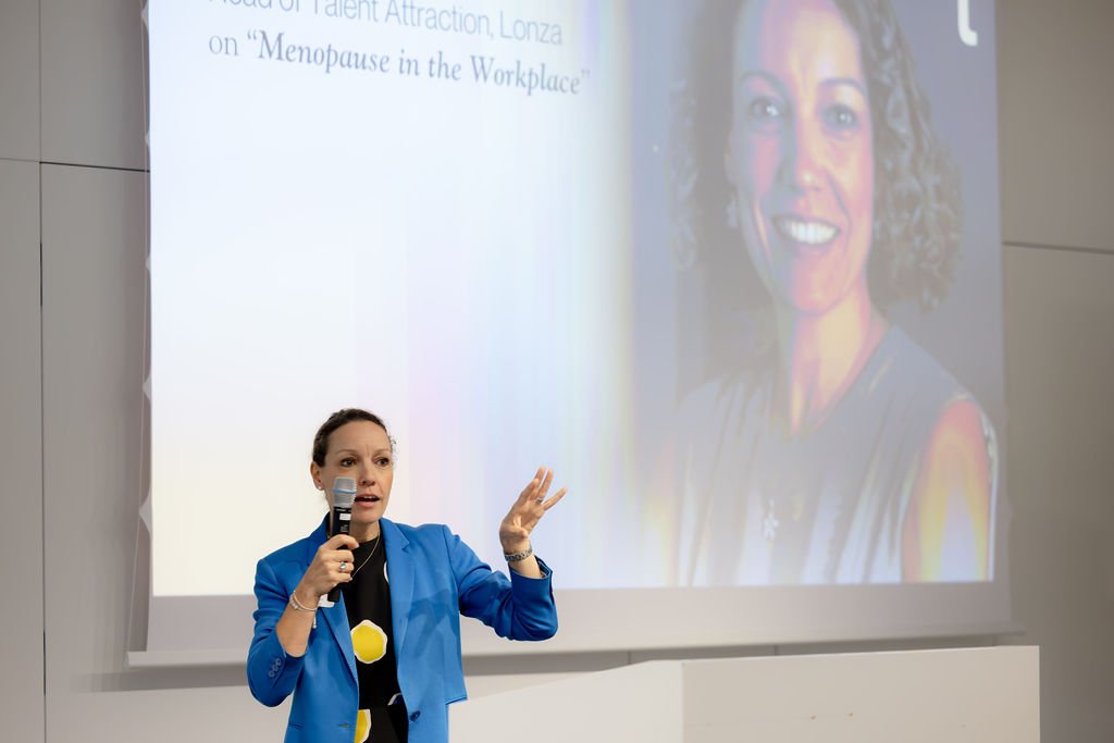 A woman in a blue blazer giving a presentation with a microphone in front of a large screen displaying a slide and a large portrait photo of herself.