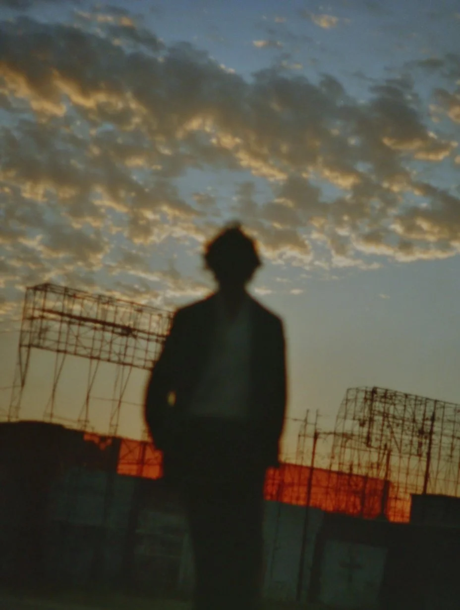 Silhouette of a person standing outdoors at sunset with a cloudy sky and light scaffolding in the background.