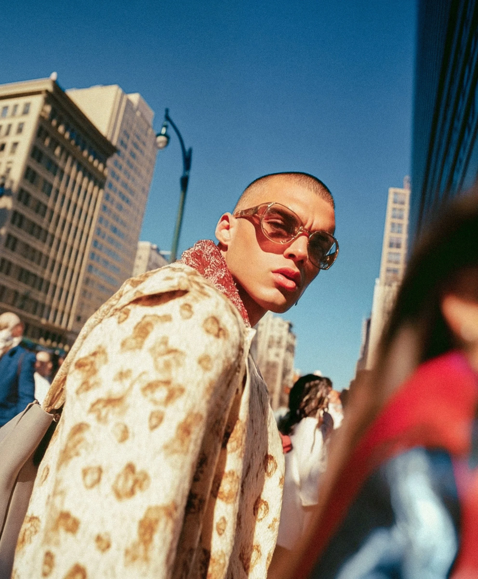 Young person with short hair wearing large sunglasses and a leopard-print coat, standing outdoors in a city with tall buildings and a clear blue sky.