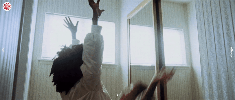 Person with curly hair raising one arm towards the ceiling in a sunlit room with large windows and wooden paneling.