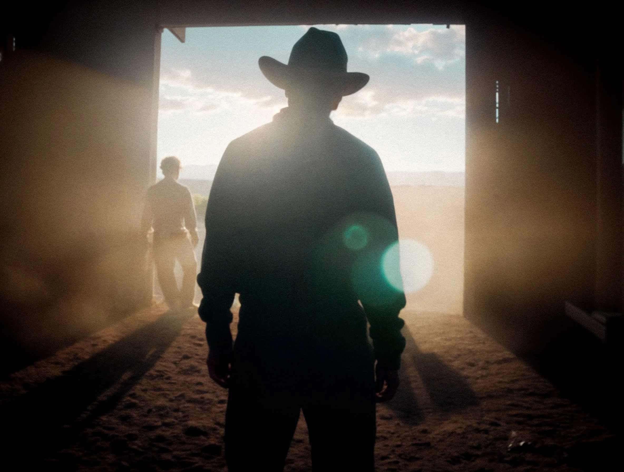 Silhouettes of two people, one wearing a cowboy hat, standing in a barn with sunlight streaming through the doorway, looking out at a rural landscape.
