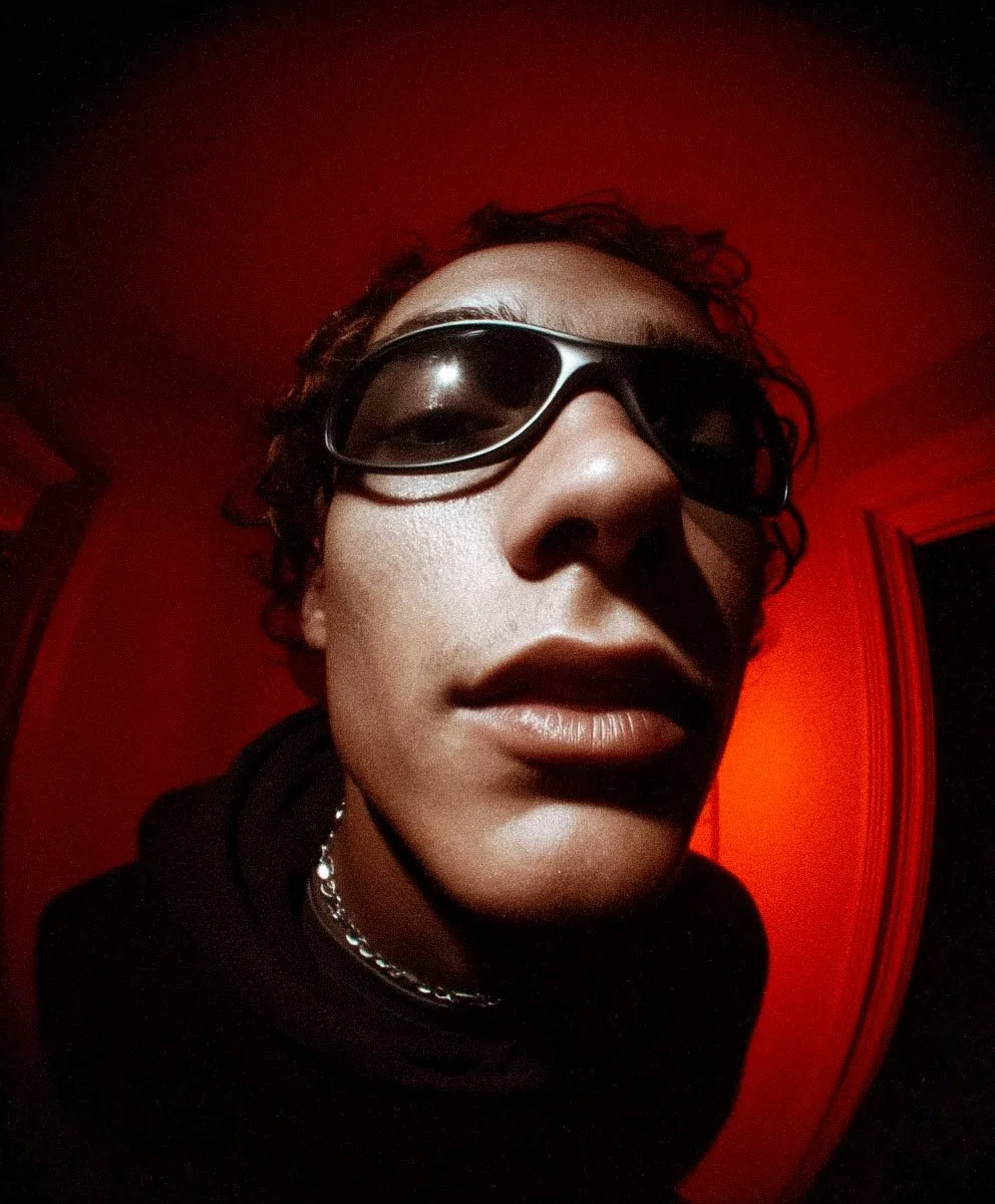 Close-up of a person wearing dark sunglasses, with a red background and curly hair.