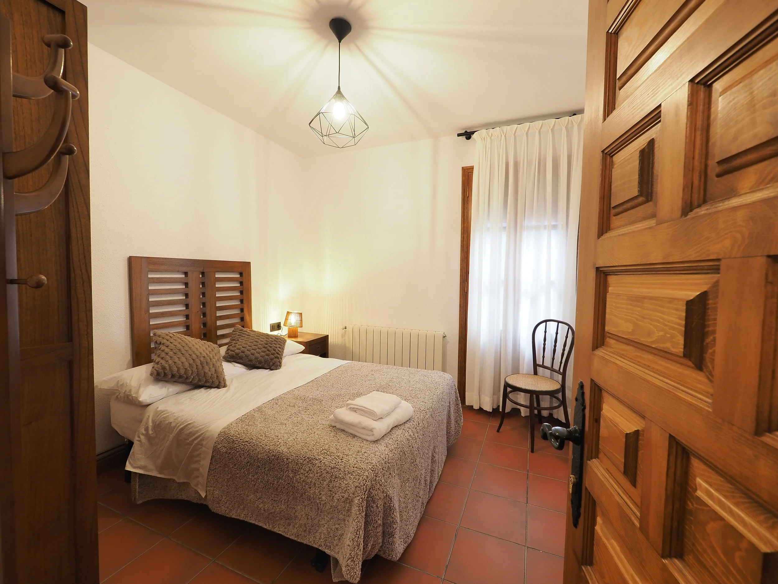 Habitación sencilla con cama de matrimonio, cabecera de madera, dos cojines color beige, lámpara de noche, cortinas blancas, silla de madera y suelo de baldosas rojas.