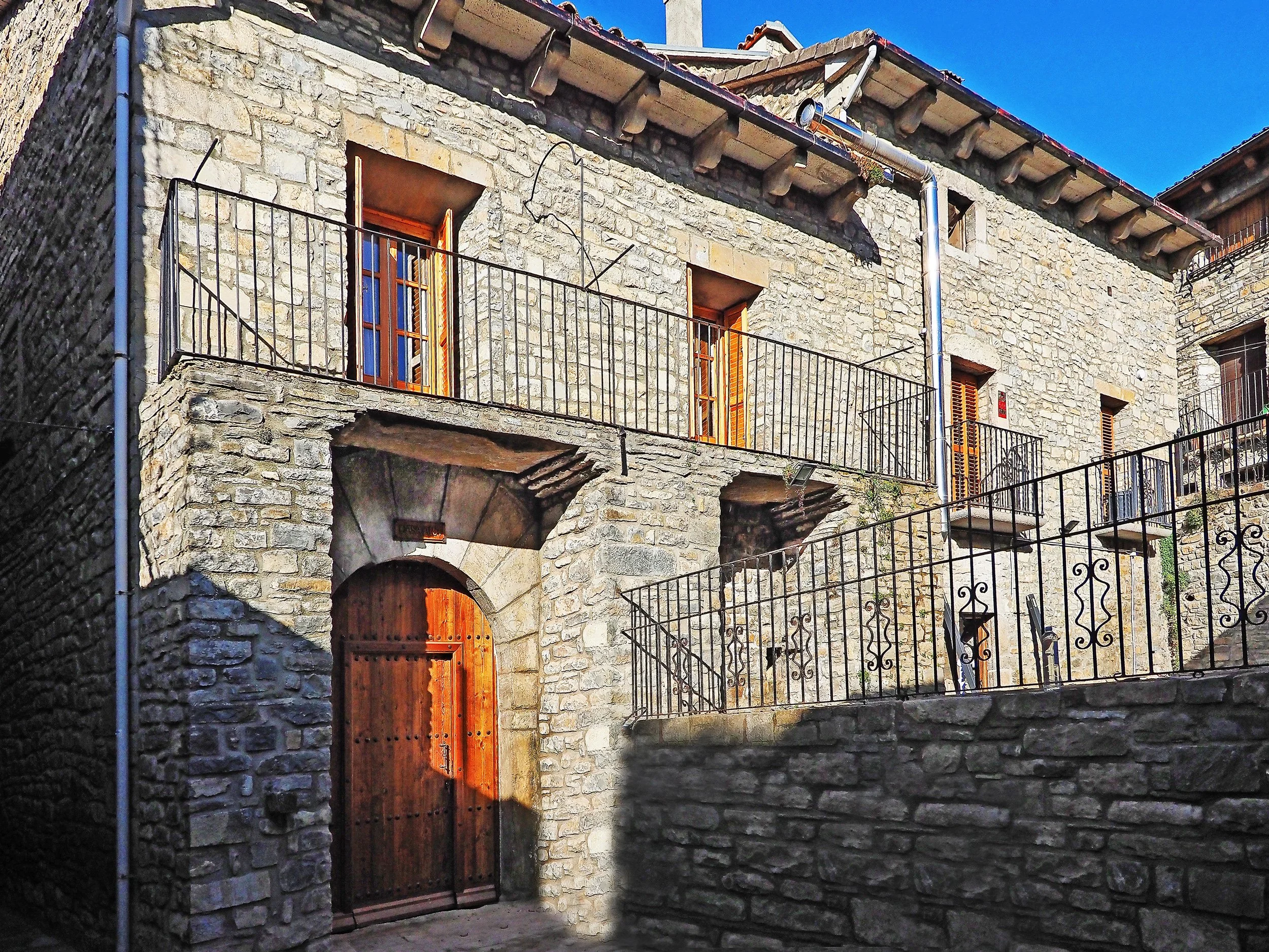 Edificio de piedra con ventanas y puertas de madera, varios balcones con barandillas de metal, en un área con escaleras y muro de piedra, bajo un cielo azul.