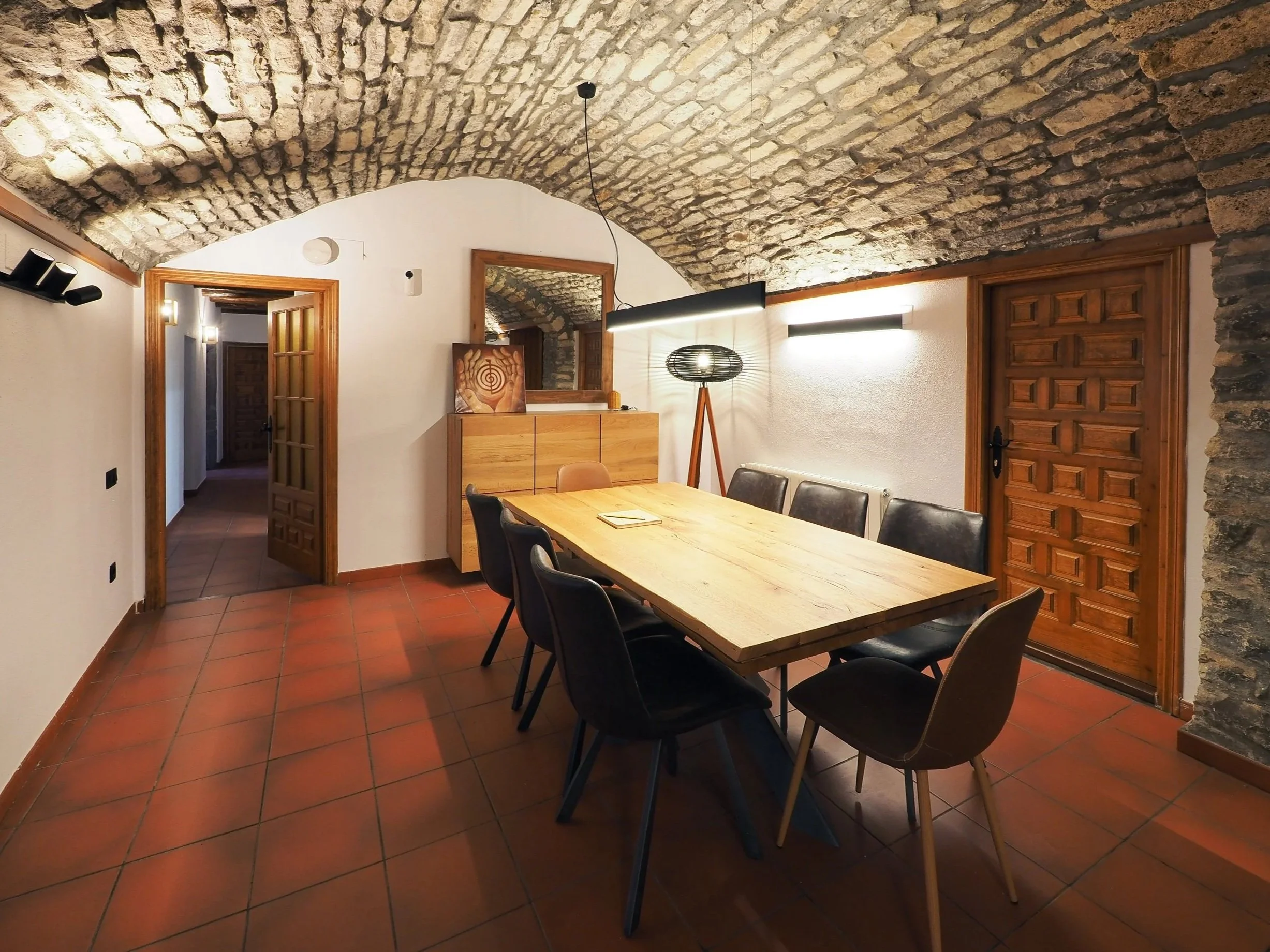 Sala de reuniones con techo de piedra, mesa de madera, sillas negras y una lámpara de pie decorativa.