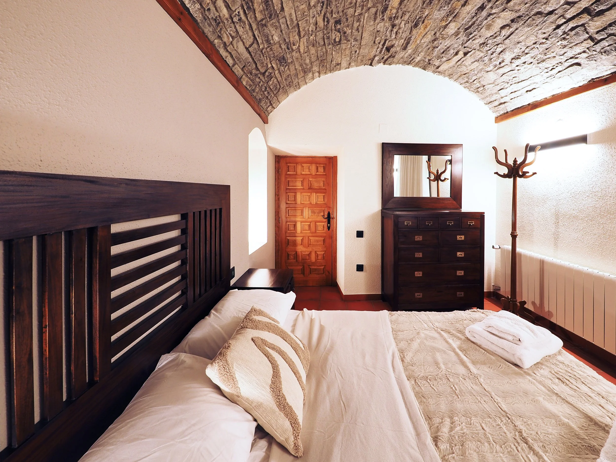 Habitación con cama de madera, cabecera con rayas, almohadas, un edredón beige, una toalla doblada en la cama, un mueble con espejo y cajones, una percha de pie y una puerta de madera