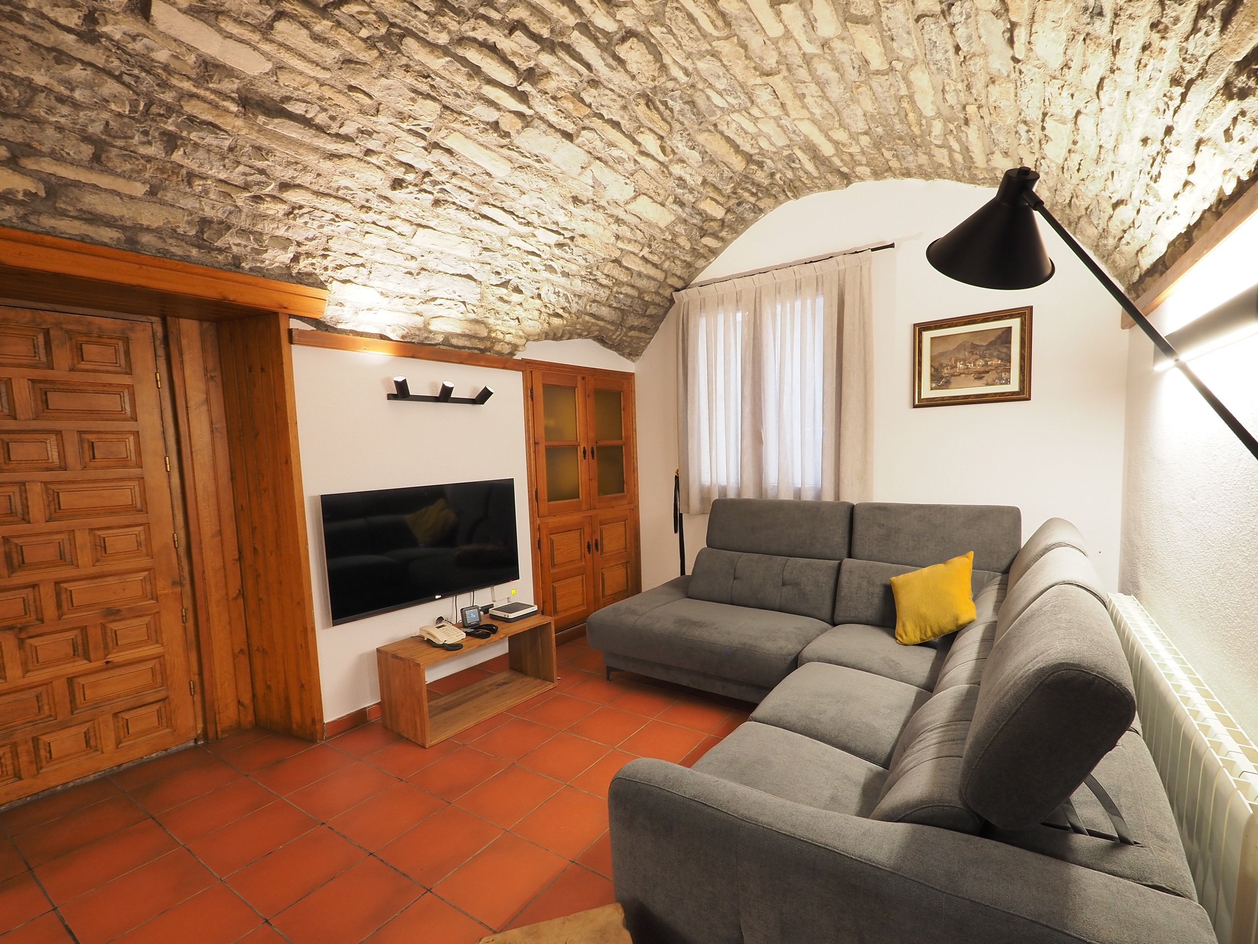 Sala de estar con paredes de piedra y techo abovedado, sofá gris con una almohada amarilla, televisión en la pared y cortinas blancas en la ventana