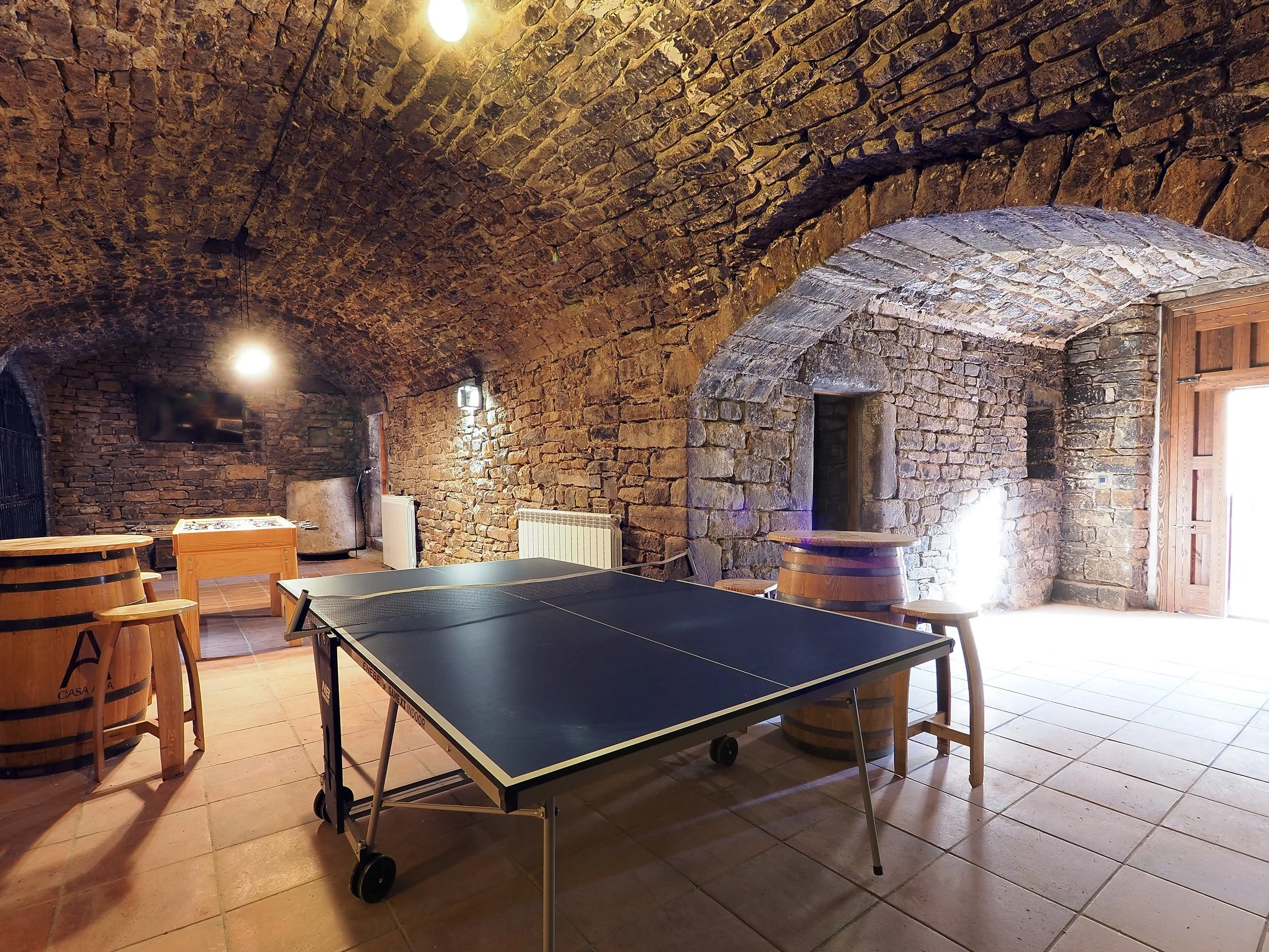 Sala con techo de arcos de piedra, mesa de ping pong, barriles de madera, televisión y puertas de madera, iluminada con luces y luz natural