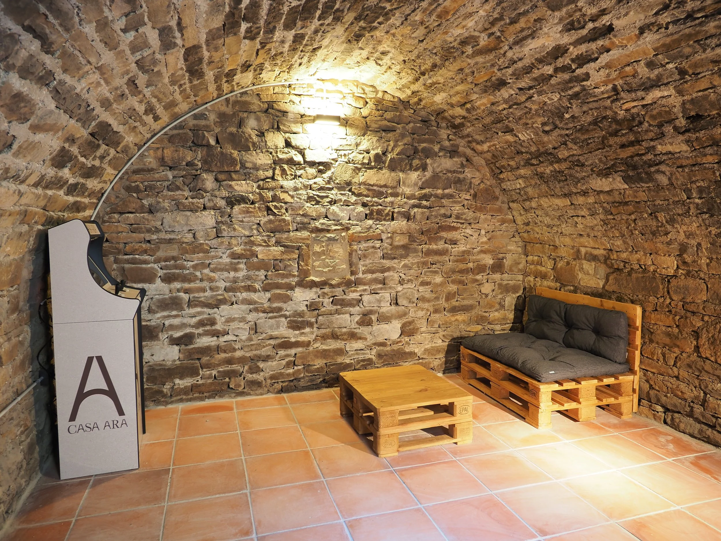 Sala de estar con paredes de piedra, suelo de baldosas, un sofá hecho con palets de madera con cojines oscuros y una mesa de madera también de palets. Hay una lámpara en arco y una estructura con la inscripción 'CASA ARA'.