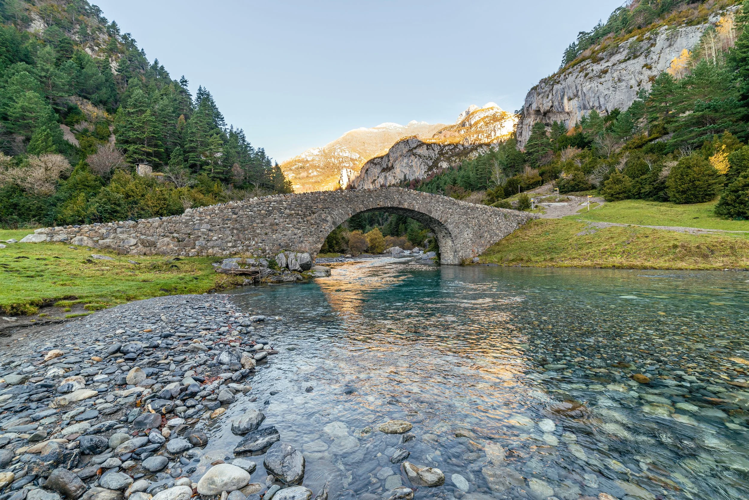 Puente de piedra sobre río en un paisaje montañoso con árboles verdes y un cielo despejado
