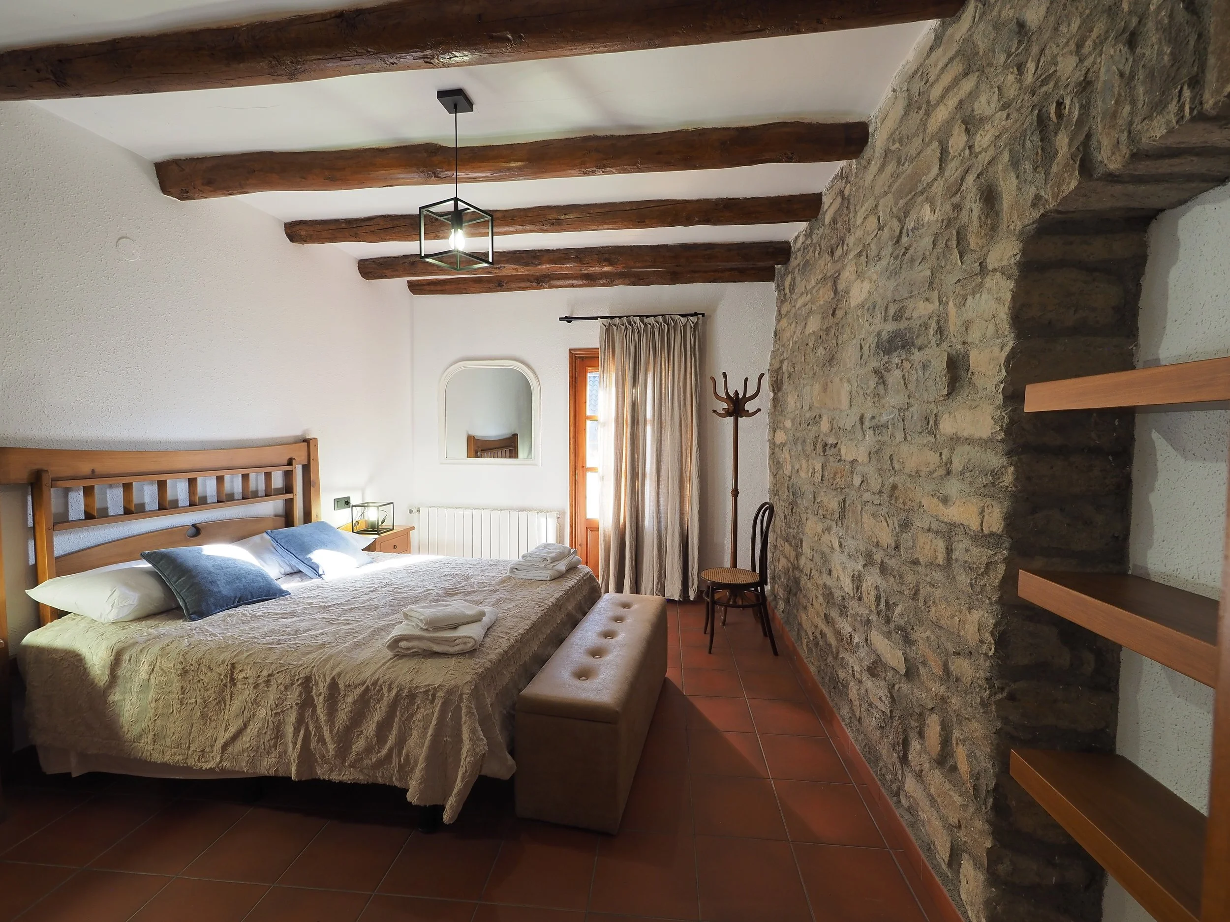 Habitación con cama, almohadas, colcha, ventana con cortinas, pared de piedra y techo con vigas de madera.