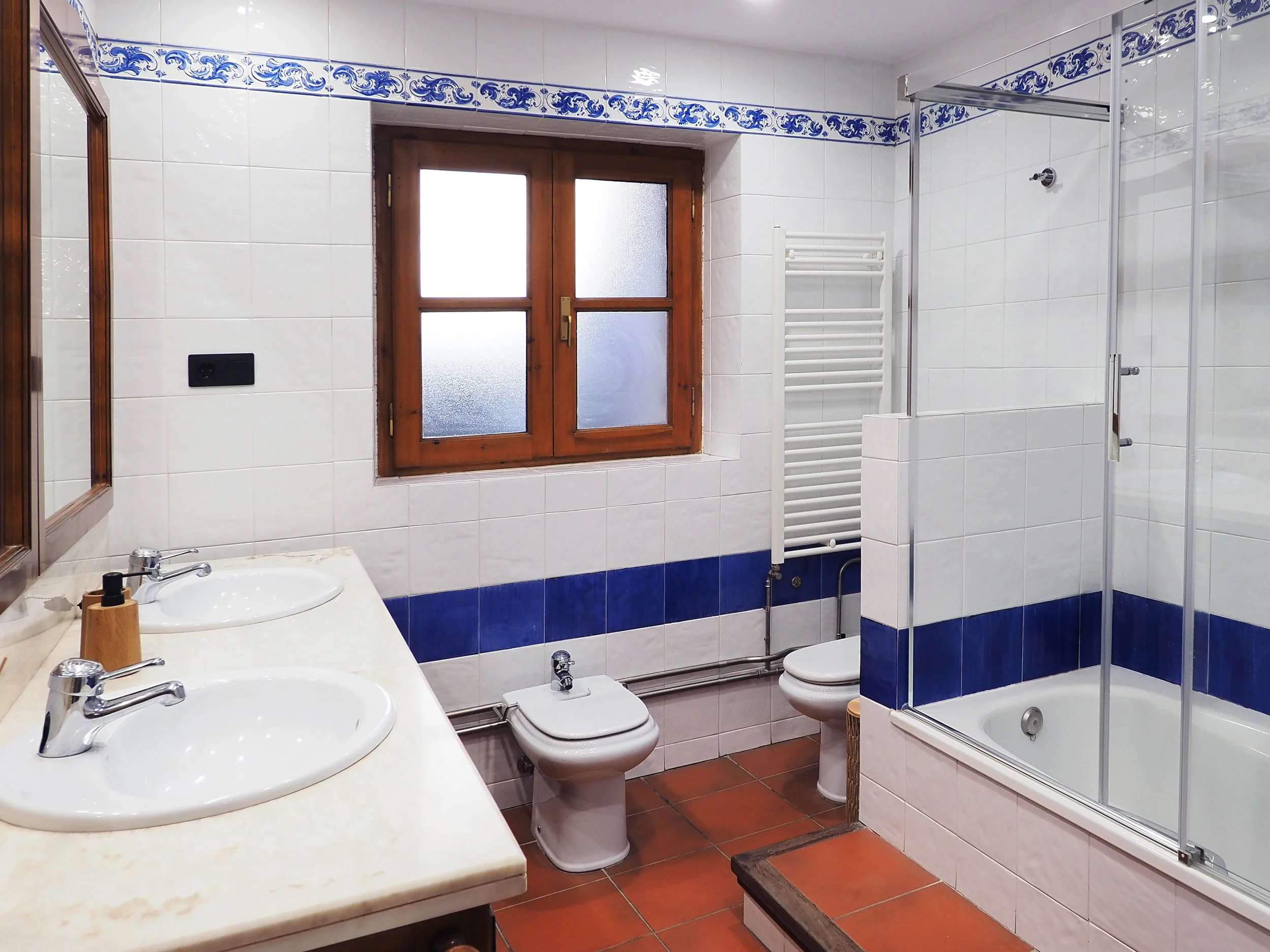 Baño con lavabo, espejo, bidé, inodoro, ducha y ventana de madera, decorado con baldosas blancas y azules.