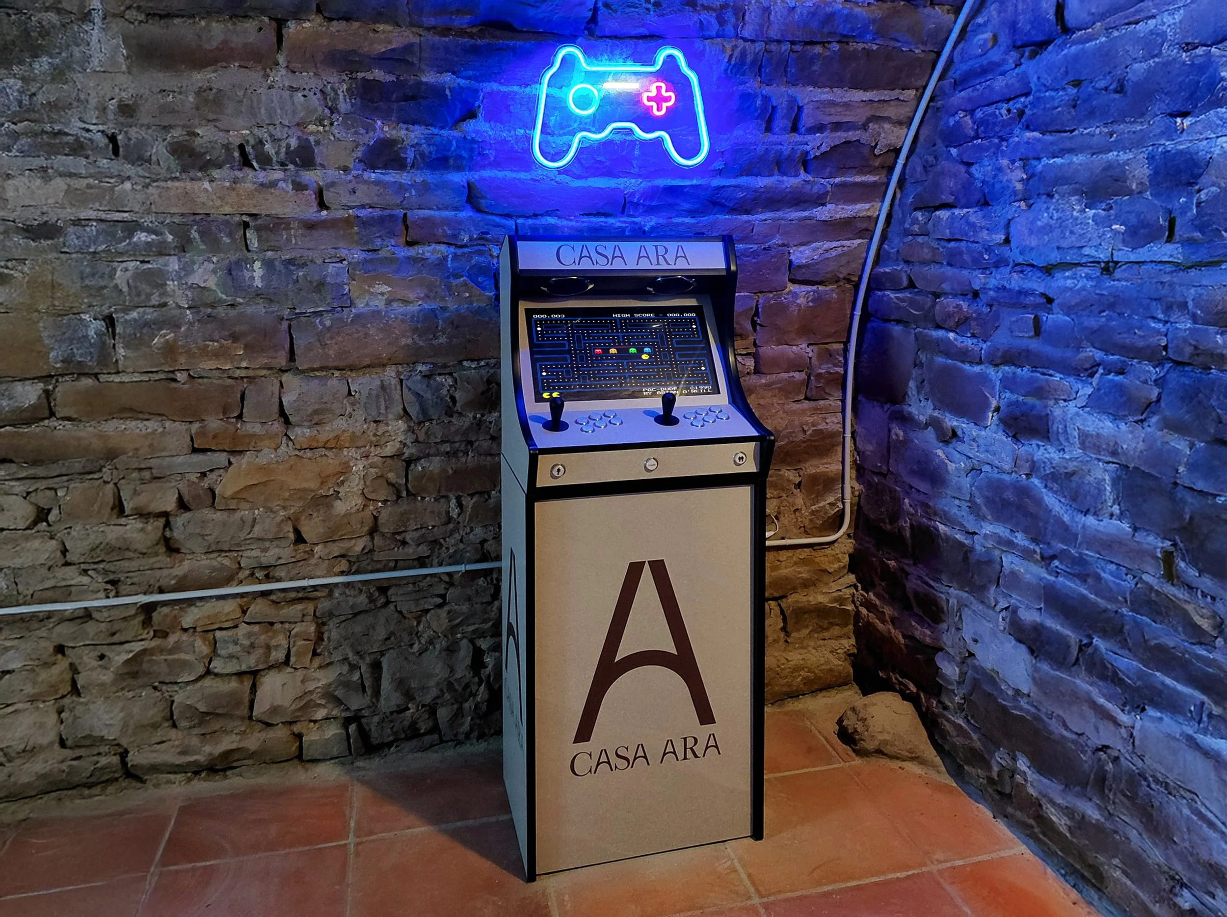 Una máquina recreativa arcade antigua con la frase 'Casa Ara' en la parte frontal, en una pared de ladrillos con letrero de neón que representa un controlador de videojuegos.
