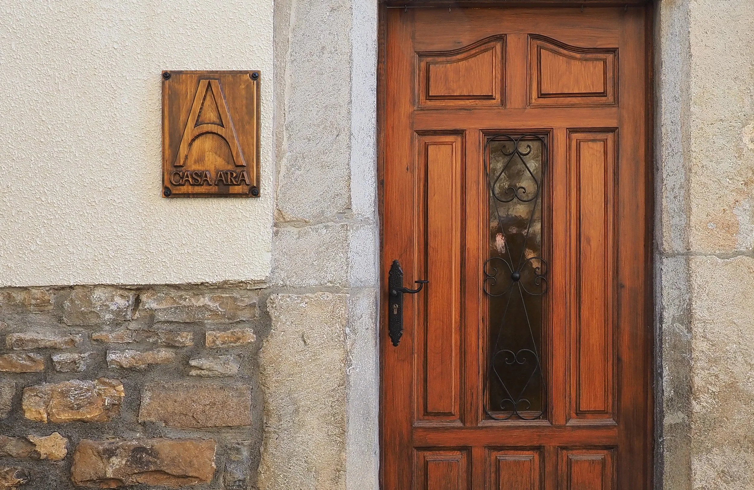 Puerta de madera con detalles de hierro, a la izquierda un cartel de madera con letras grabadas que dice "Casa Ara", paredes de piedra y estuco en el fondo.