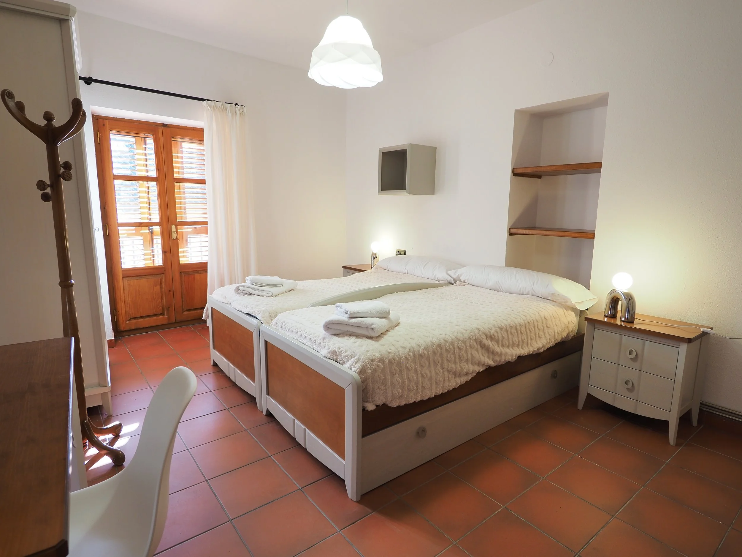 Habitación con dos camas, una junto a la otra, con sábanas blancas y toallas dobladas, mesita de noche a la derecha con lámpara y tetera, paredes blancas, ventana de madera con cortinas blancas, armario para colgar en la esquina izquierda, azulejos t