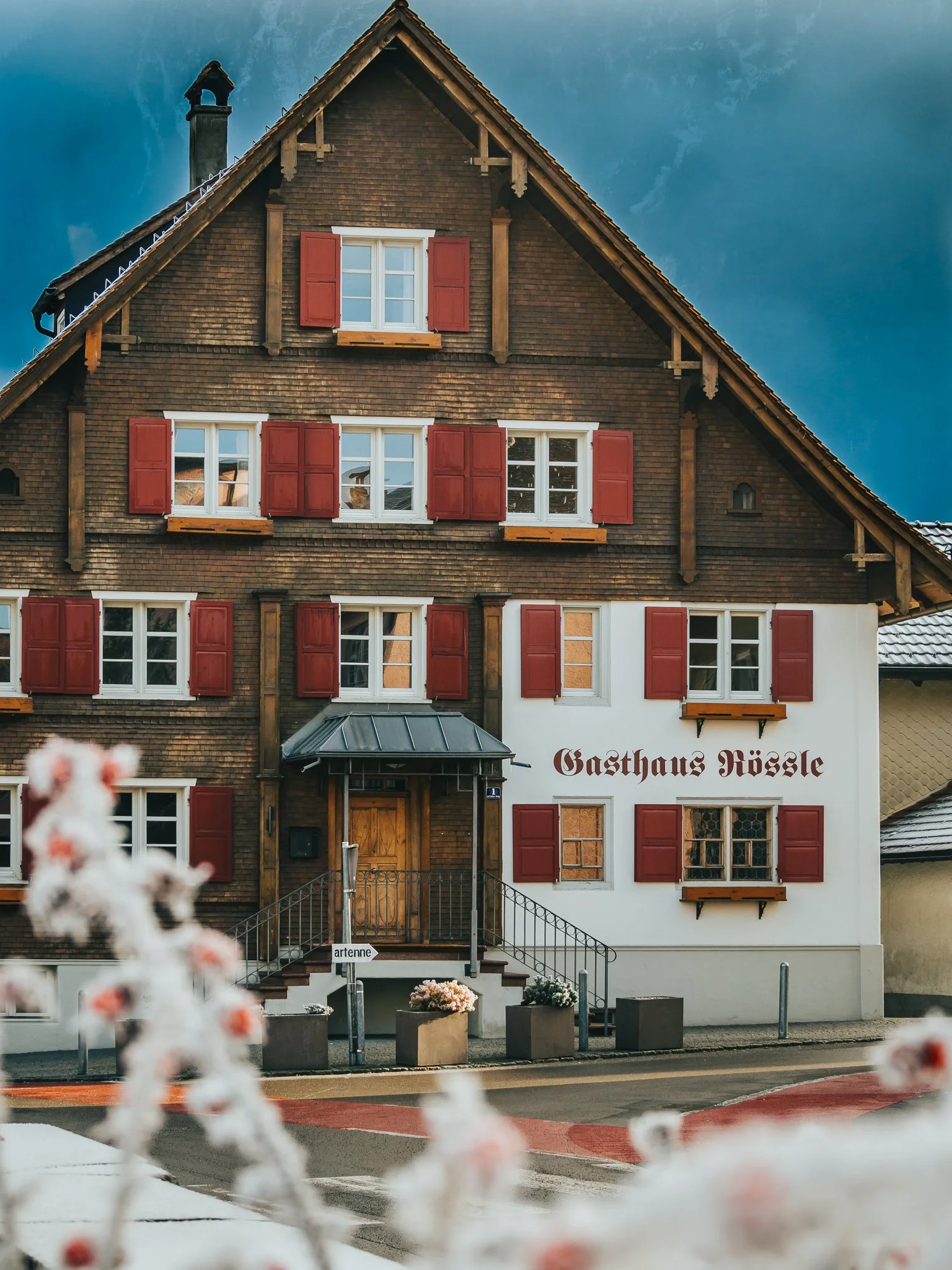 Ein traditionelles Gasthaus mit mehrstöckigem Gebäude, braunen Holzschindeln, roten Fensterläden und einem Eingang mit Treppe, umgeben von blühenden Blumen.