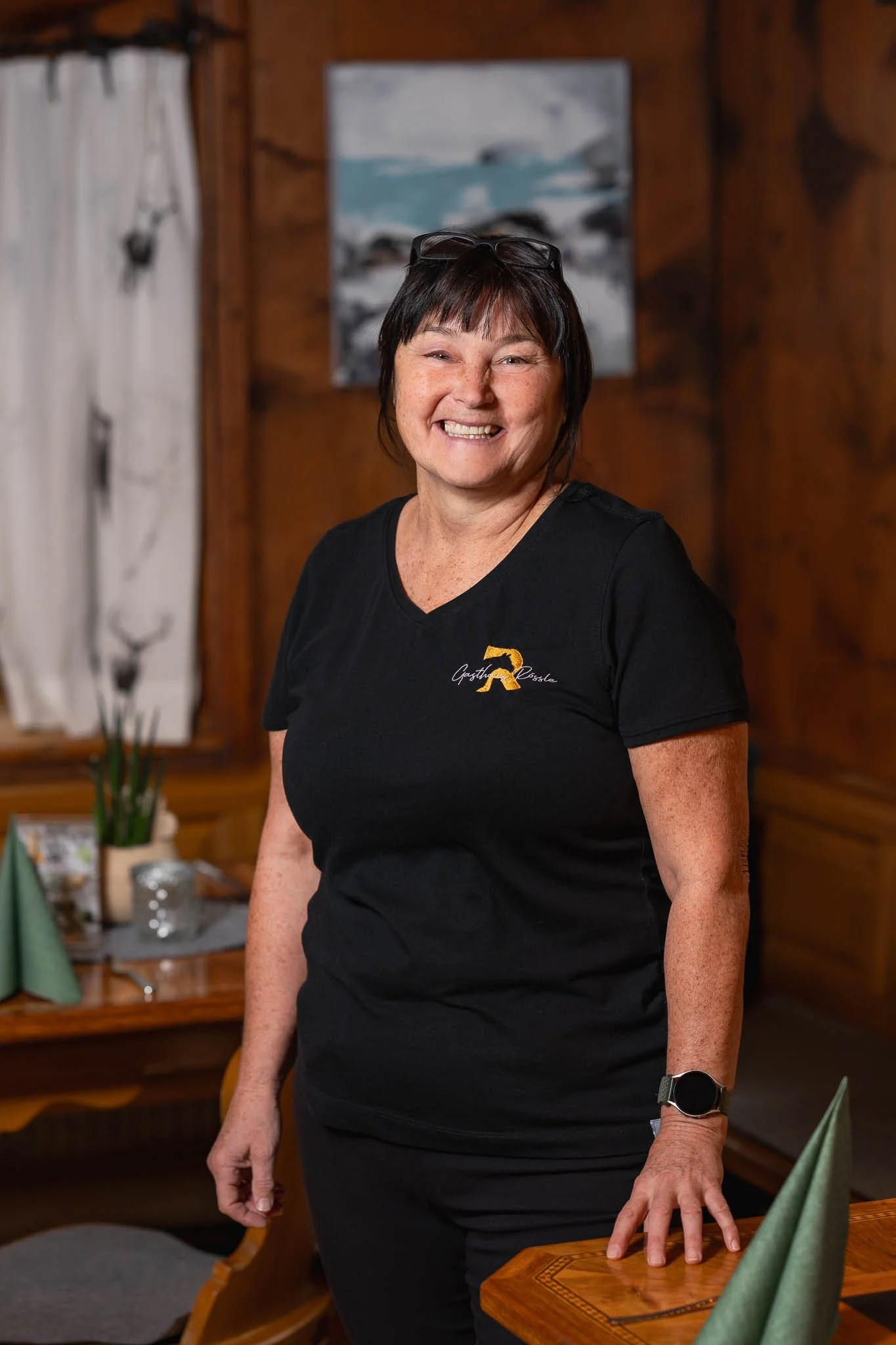 Eine lachende Frau in einem schwarzen T-Shirt mit einem Logo, die in einem rustikalen Restaurant sitzt.