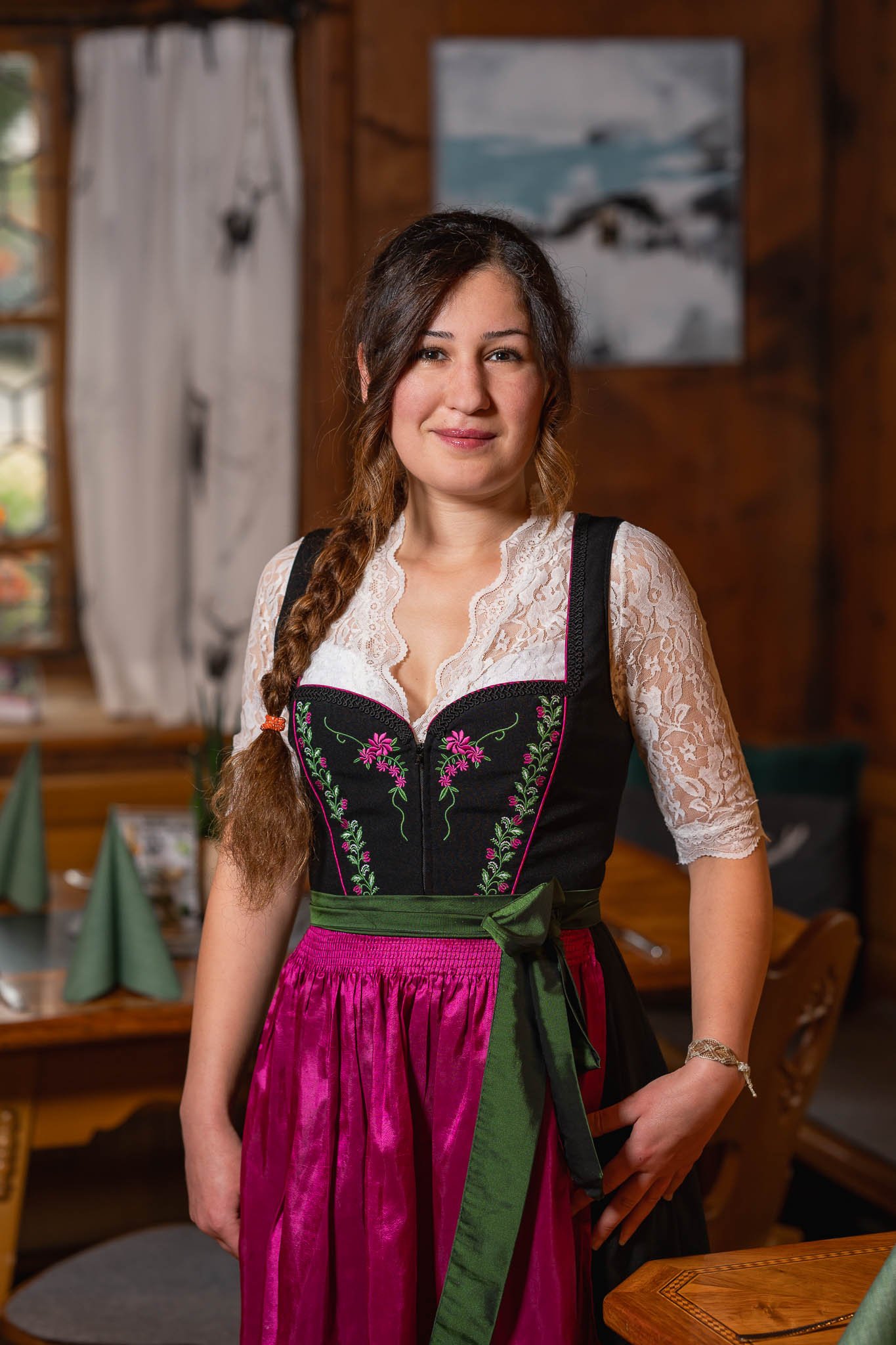Junge Frau in traditioneller Tracht in einem Holzrestaurant, mit kurzer Lockenfrisur, stehend hinter einem Tisch, lächelnd, mit einer bunten Dekoration im Hintergrund.