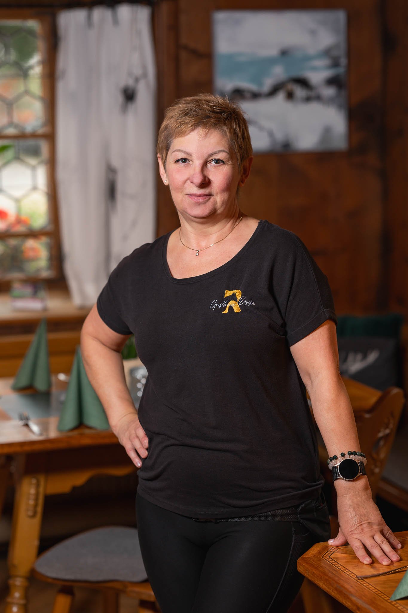 Eine Frau steht in einem rustikalen Restaurant mit Holzinterieur, trägt ein schwarzes T-Shirt mit einem Logo, rechte Hand auf einem Tisch, blickt in die Kamera.