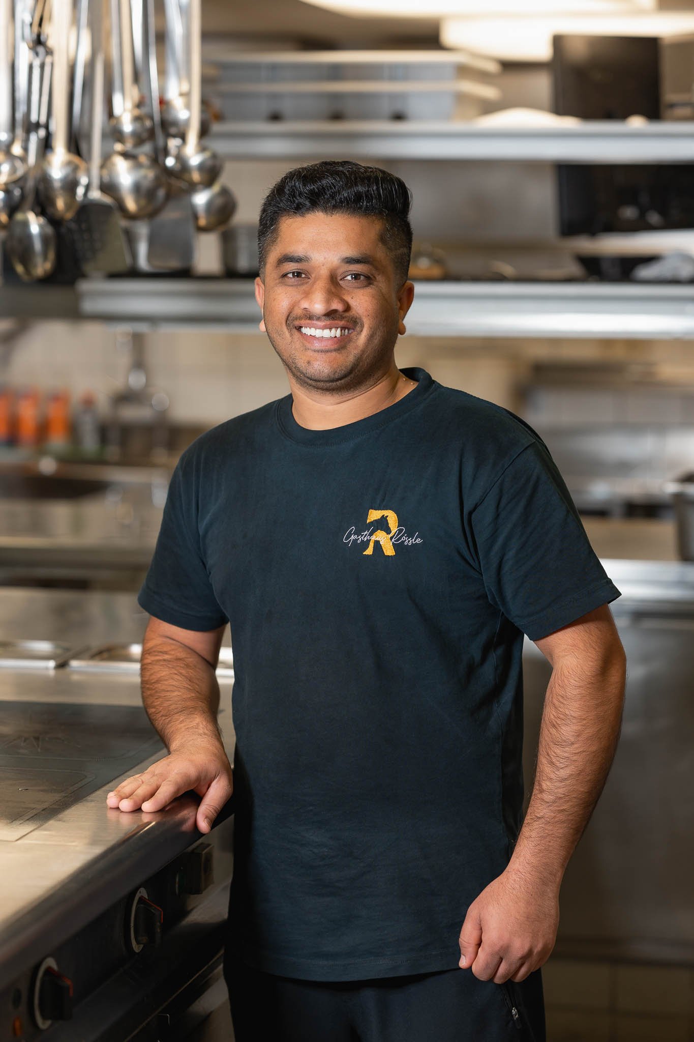 Ein lachender Mann in einer schwarzen T-Shirt steht in einer professionellen Küche mit Metallutensilien im Hintergrund.