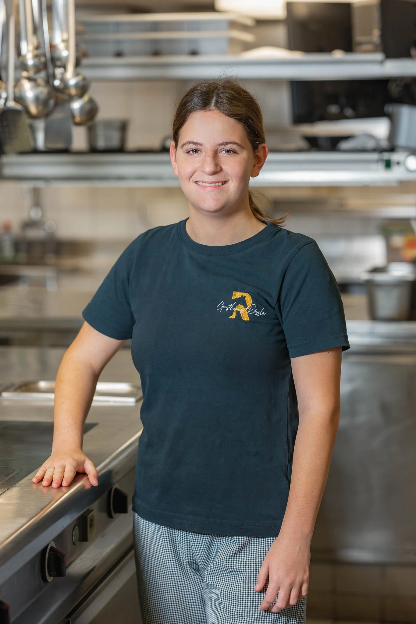 Eine junge Frau in einer schwarzen T-Shirt mit einem aufgestickten goldfarbenen D und weißen karierten Hosen steht in einer professionellen Küche, die im Hintergrund mit Kochutensilien und Kochgeräten ausgestattet ist.