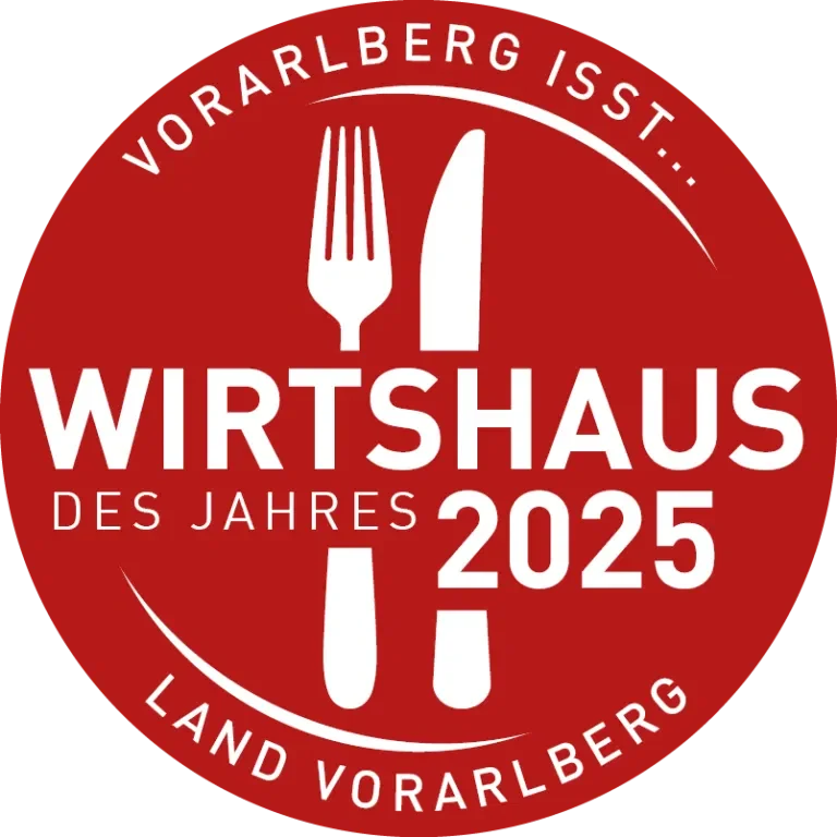 Rundes Logo mit rotem Hintergrund, weißen Texten und einem Messer und einer Gabel in der Mitte. Der Text lautet: 'Vorarlberg ist... Wirtshaus des Jahres 2025, Land Vorarlberg'.