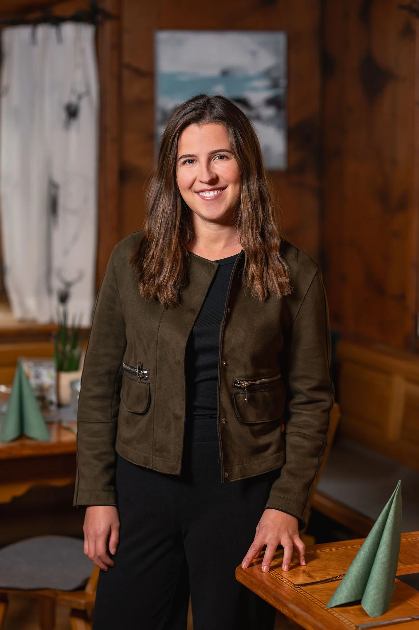 Lächelnde Frau mit braunen Haaren in einem dunklen Raum mit Holzwänden, auf einem Tisch stehen grüne Stoffservietten.