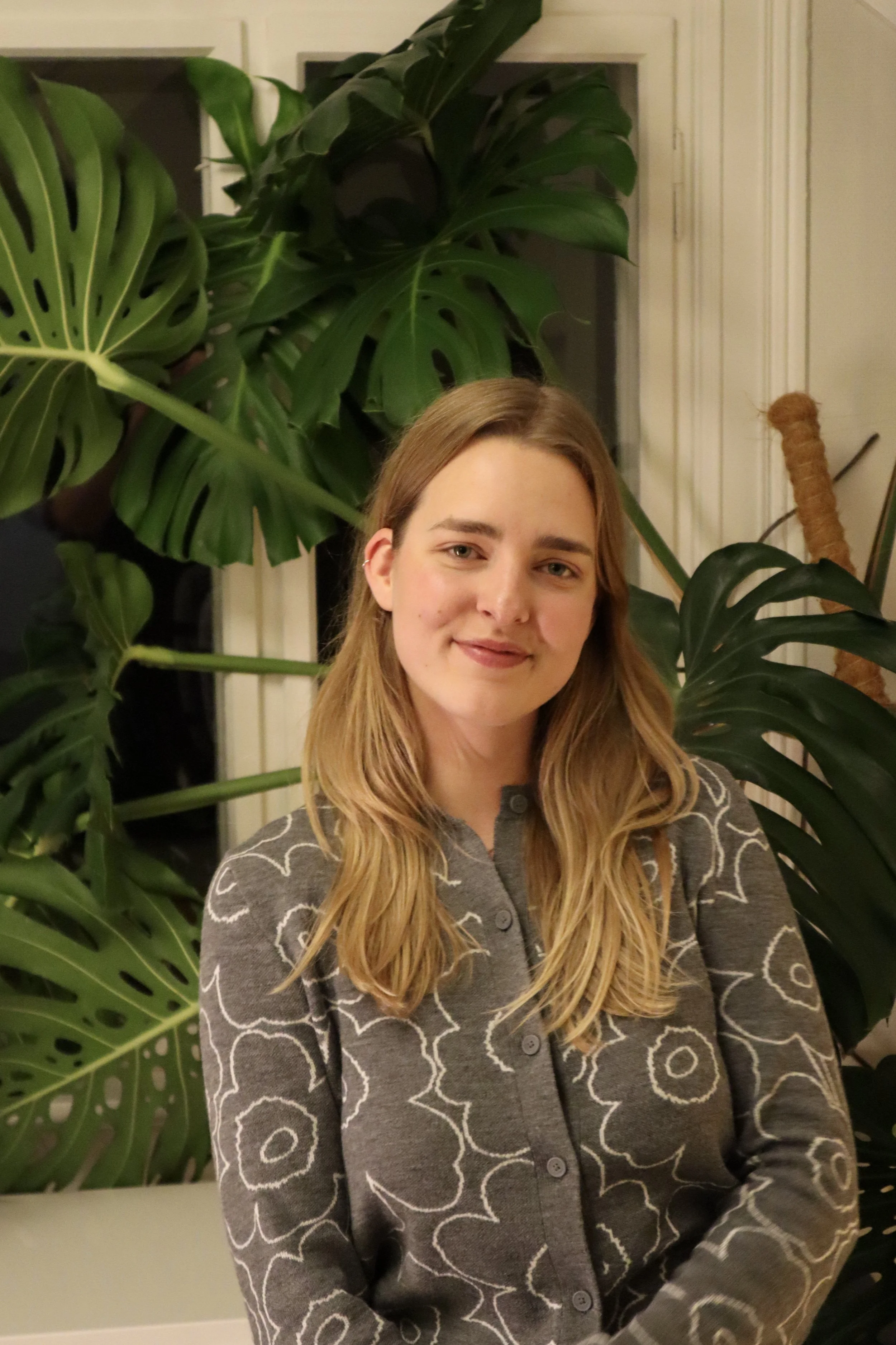 A young woman with long blonde hair smiling in front of large green monstera plants inside a room.