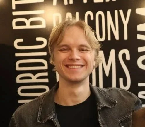 Smiling young man with blonde hair wearing a black jacket, standing in front of a black and white wall with words.