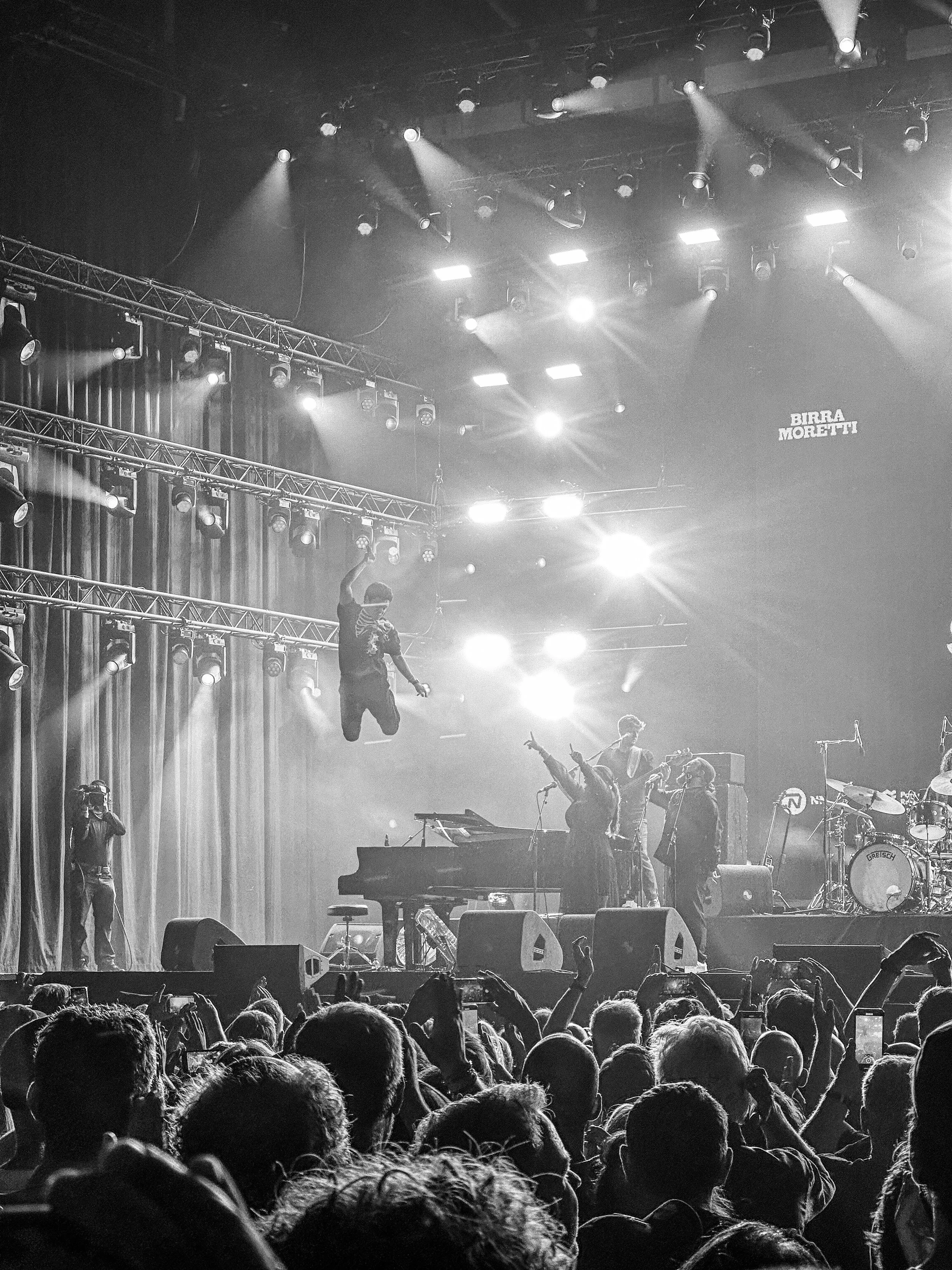 Black and white photo of a concert stage with musicians performing, several spotlights shining down, a vocalist jumping in the air, a crowd in the foreground, and the sign 'Birra Moretti' visible in the background.