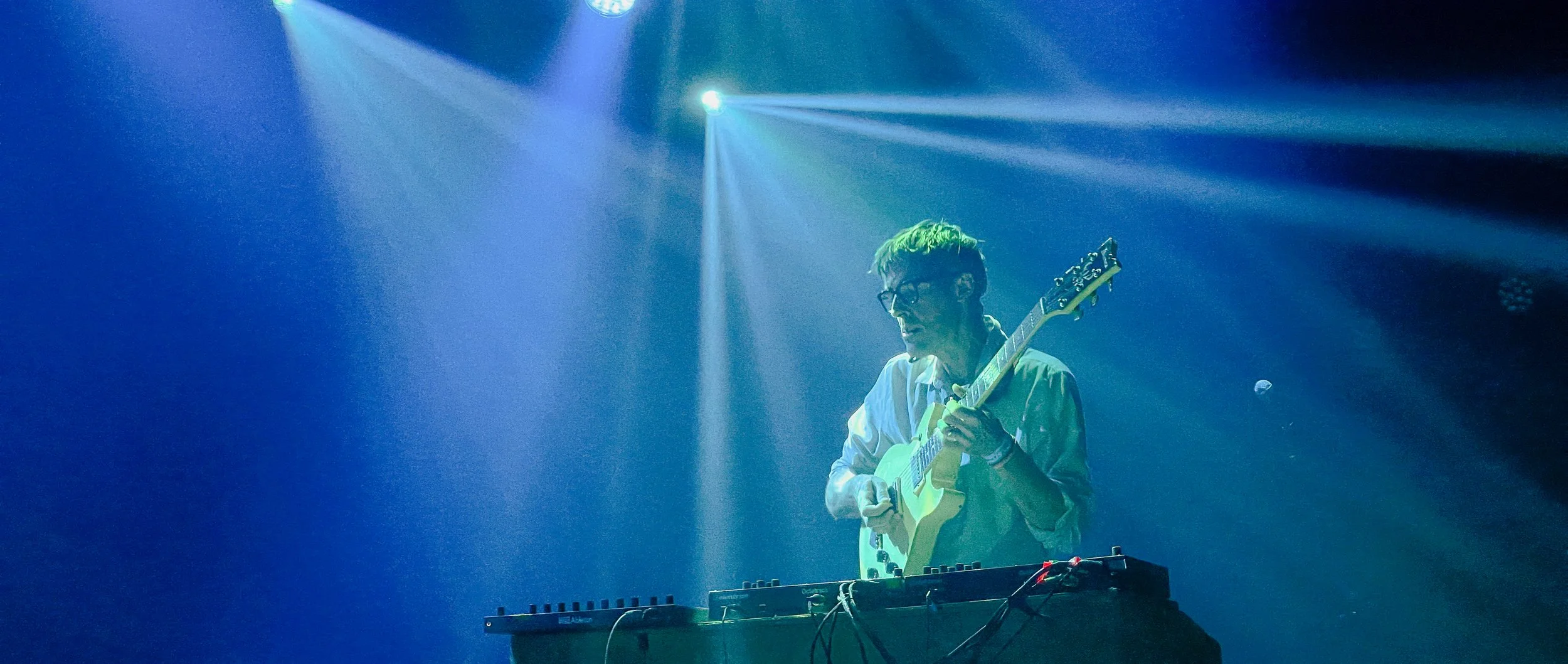 A musician with green hair and glasses playing an electric guitar on stage, with colorful stage lights shining down.