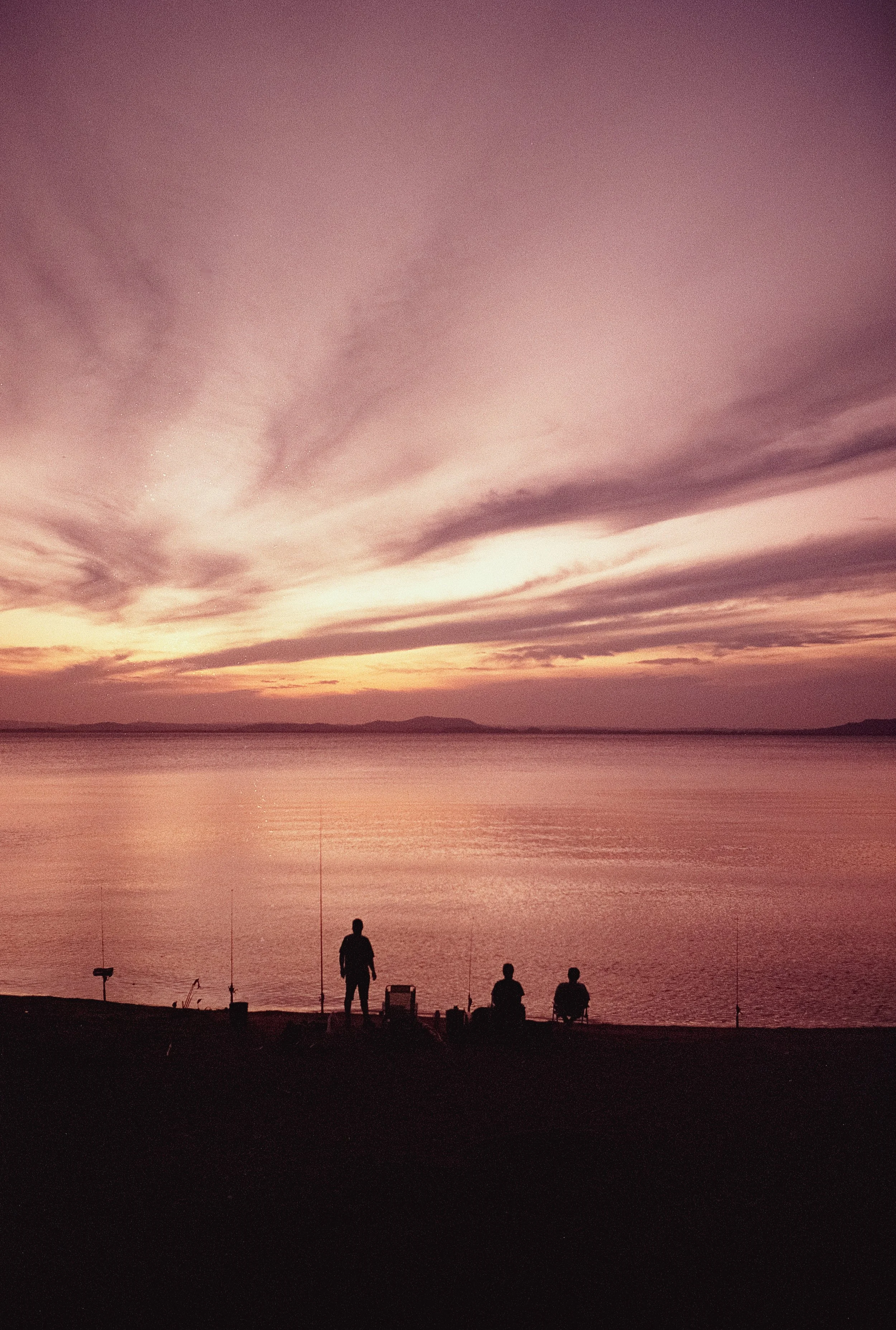 A sunset over a body of water with a colorful sky, silhouettes of three people and fishing gear on the shore.